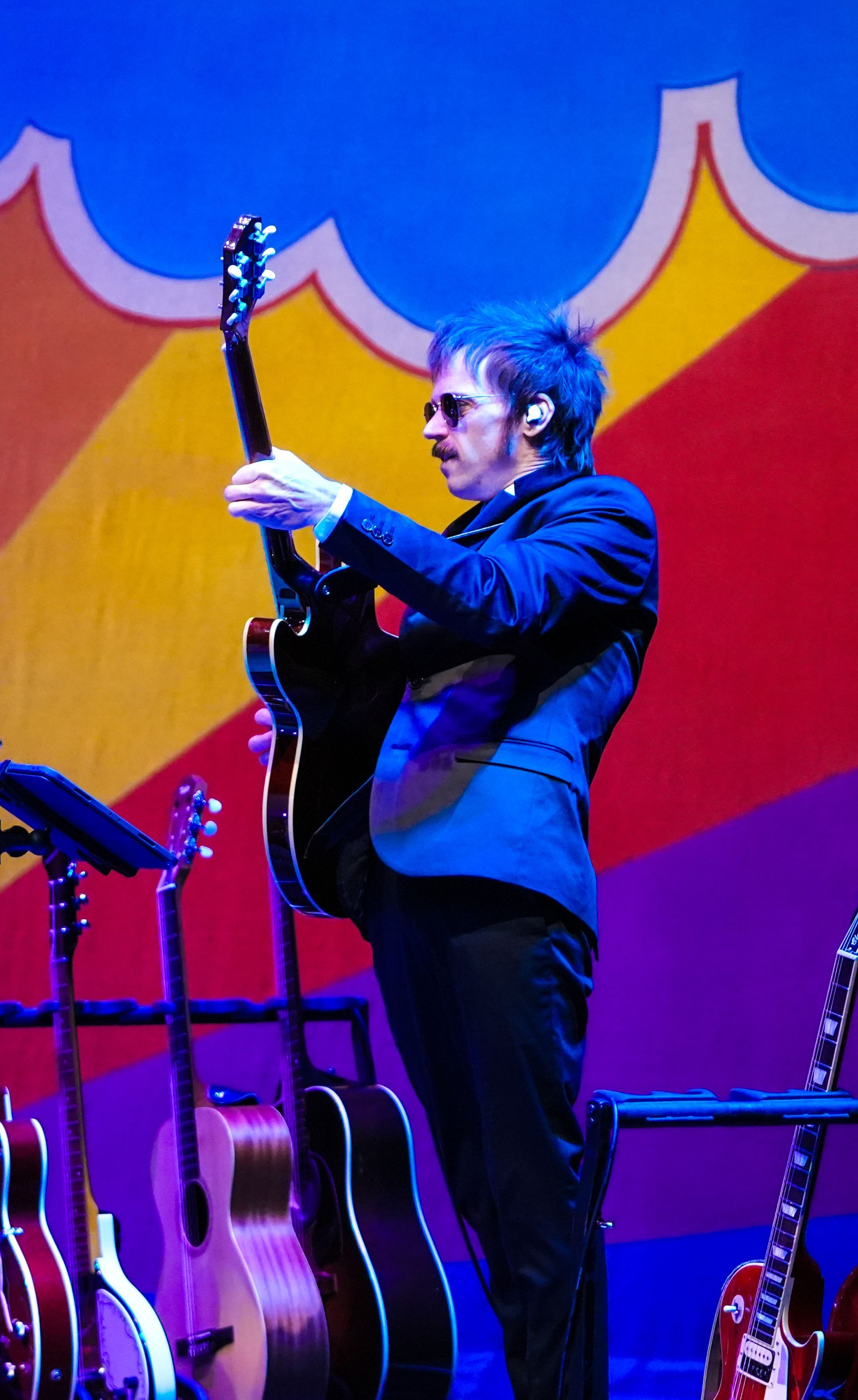 Man in dark suit holding a black guitar on stage with colorful backdrop and other guitars.