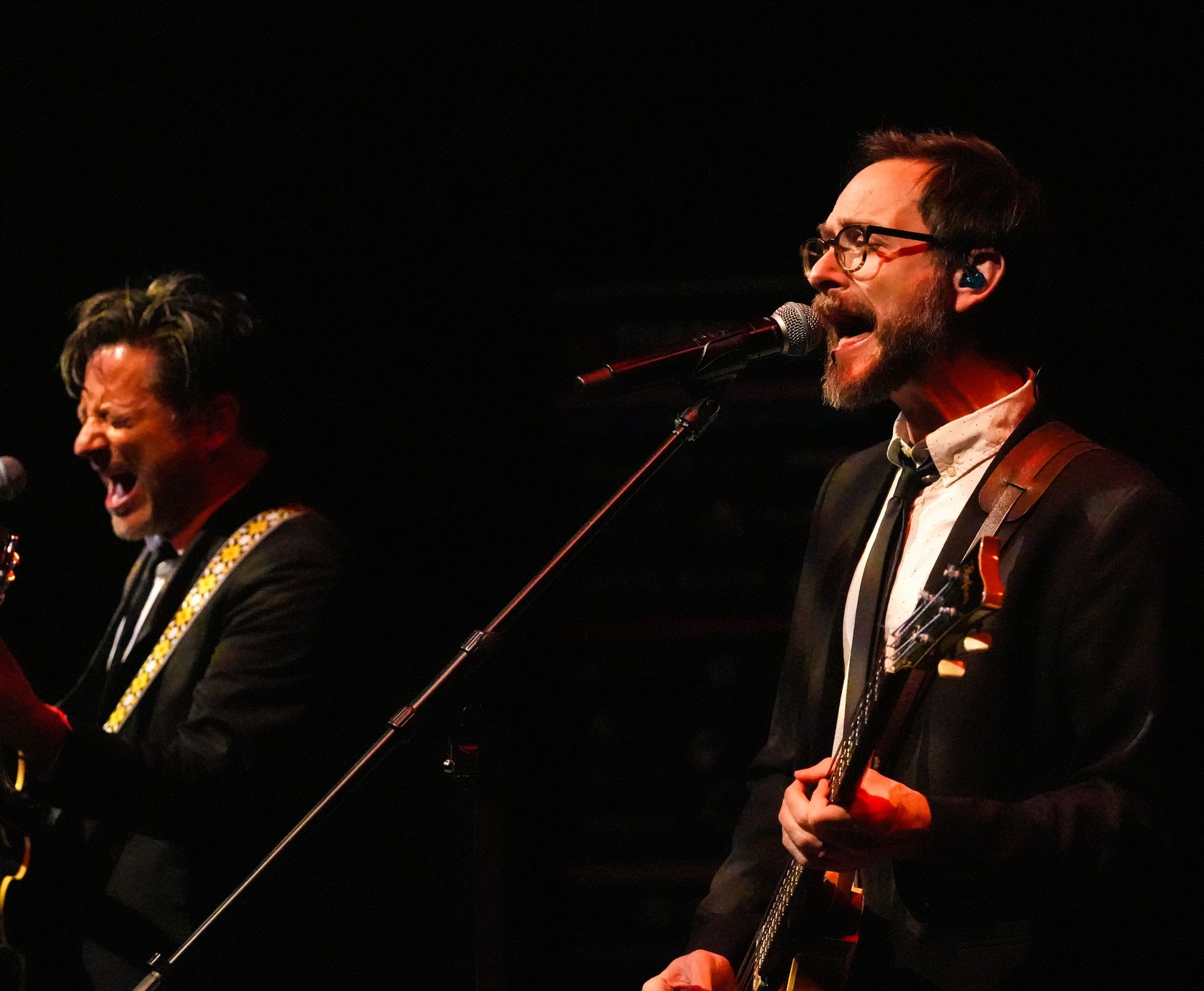 Two men singing and playing guitars on stage, illuminated by stage lights. One man has glasses and holds a guitar.