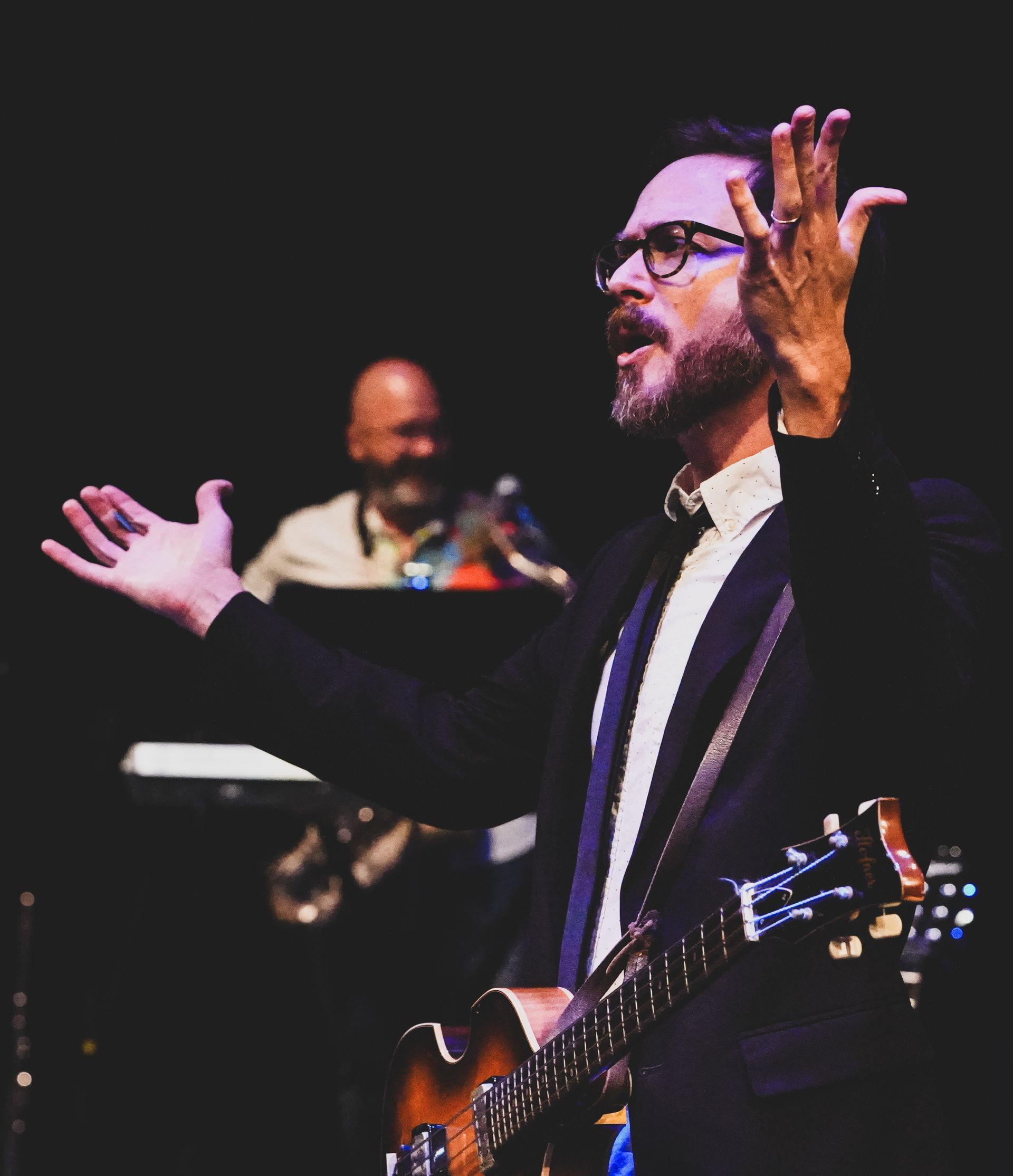 Man in suit singing and playing guitar on stage, arms raised.