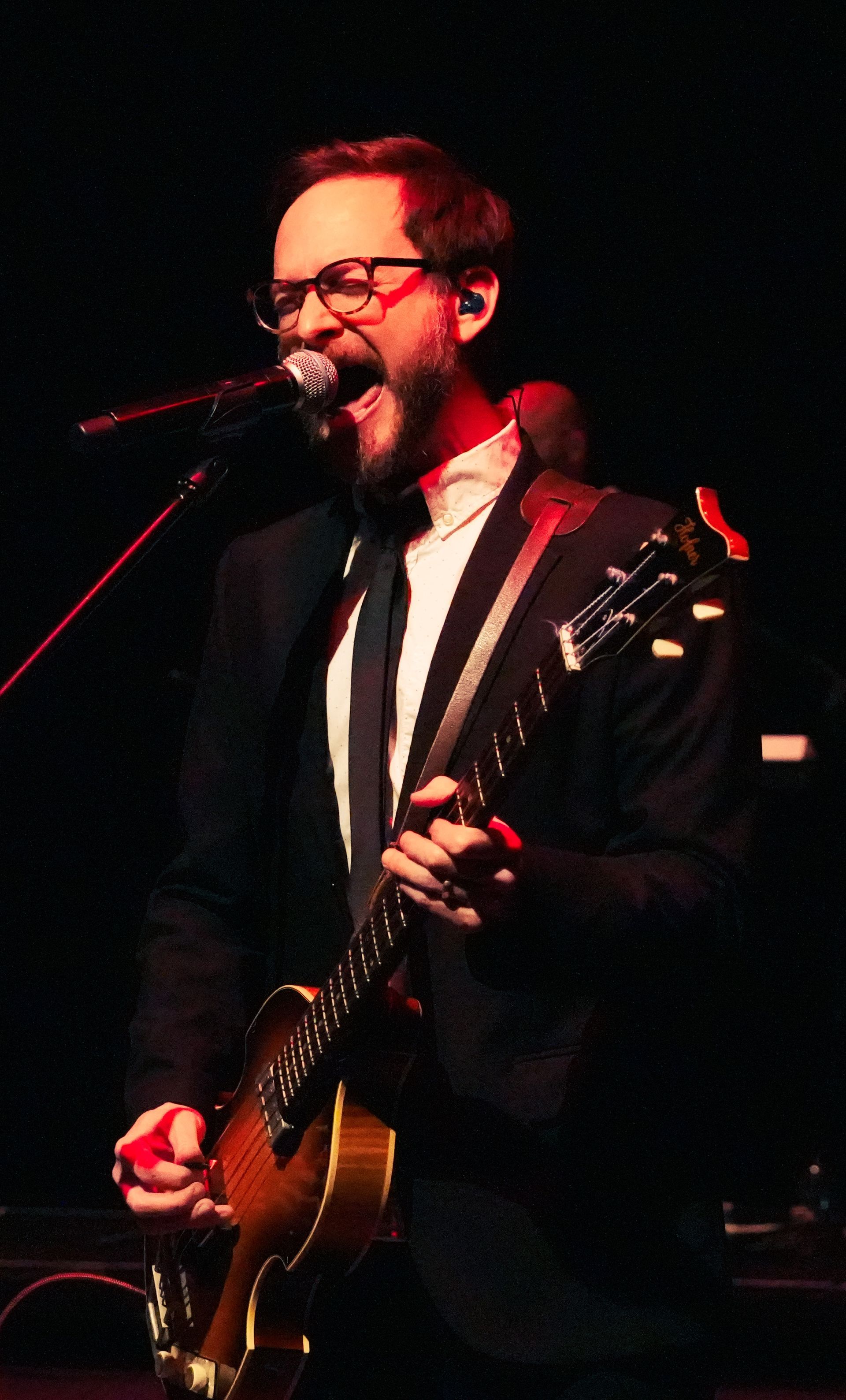 Man in glasses sings and plays guitar on stage, wearing a suit and tie.