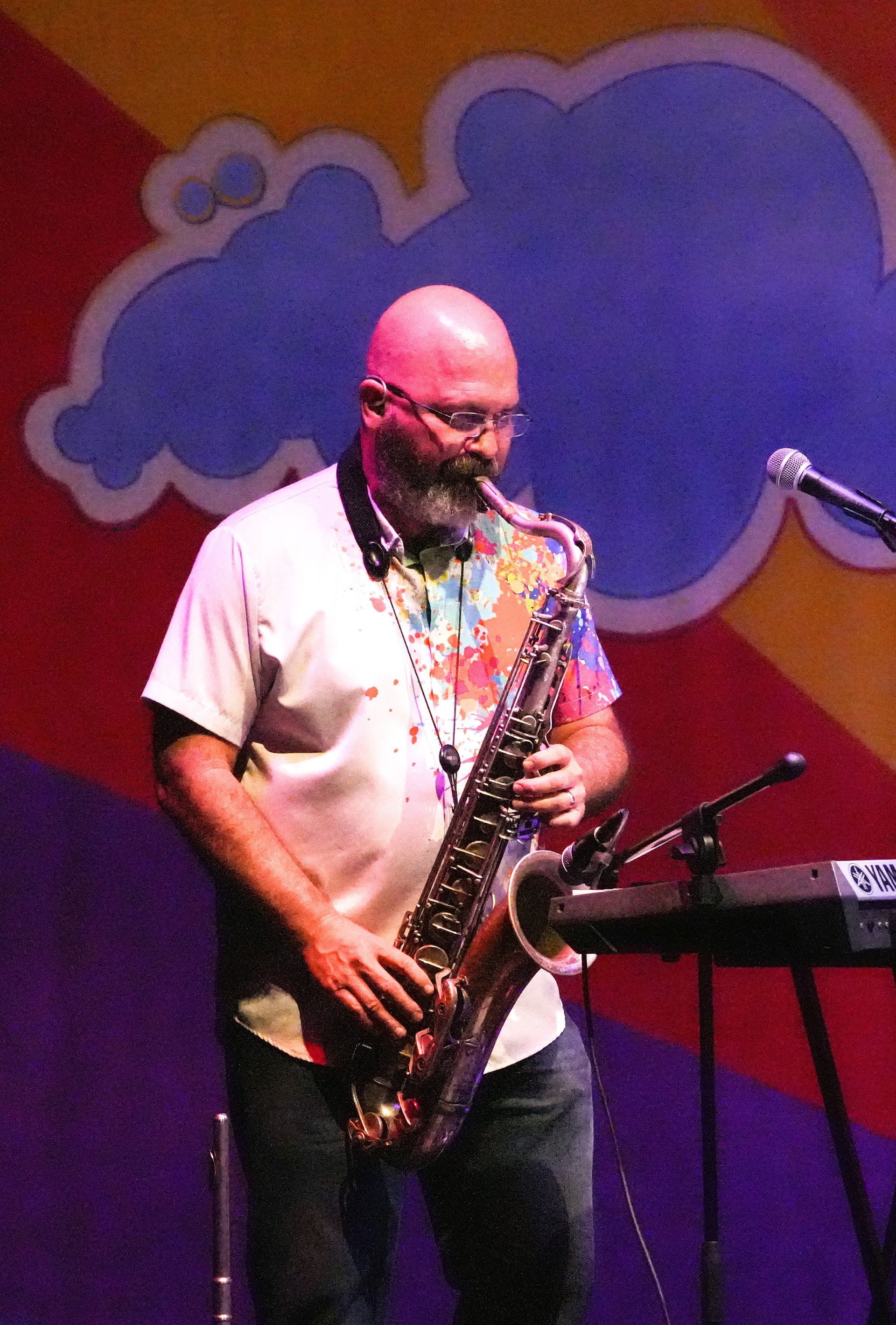 Man playing a saxophone on stage, wearing a tie-dye shirt. A colorful cloud backdrop is visible.