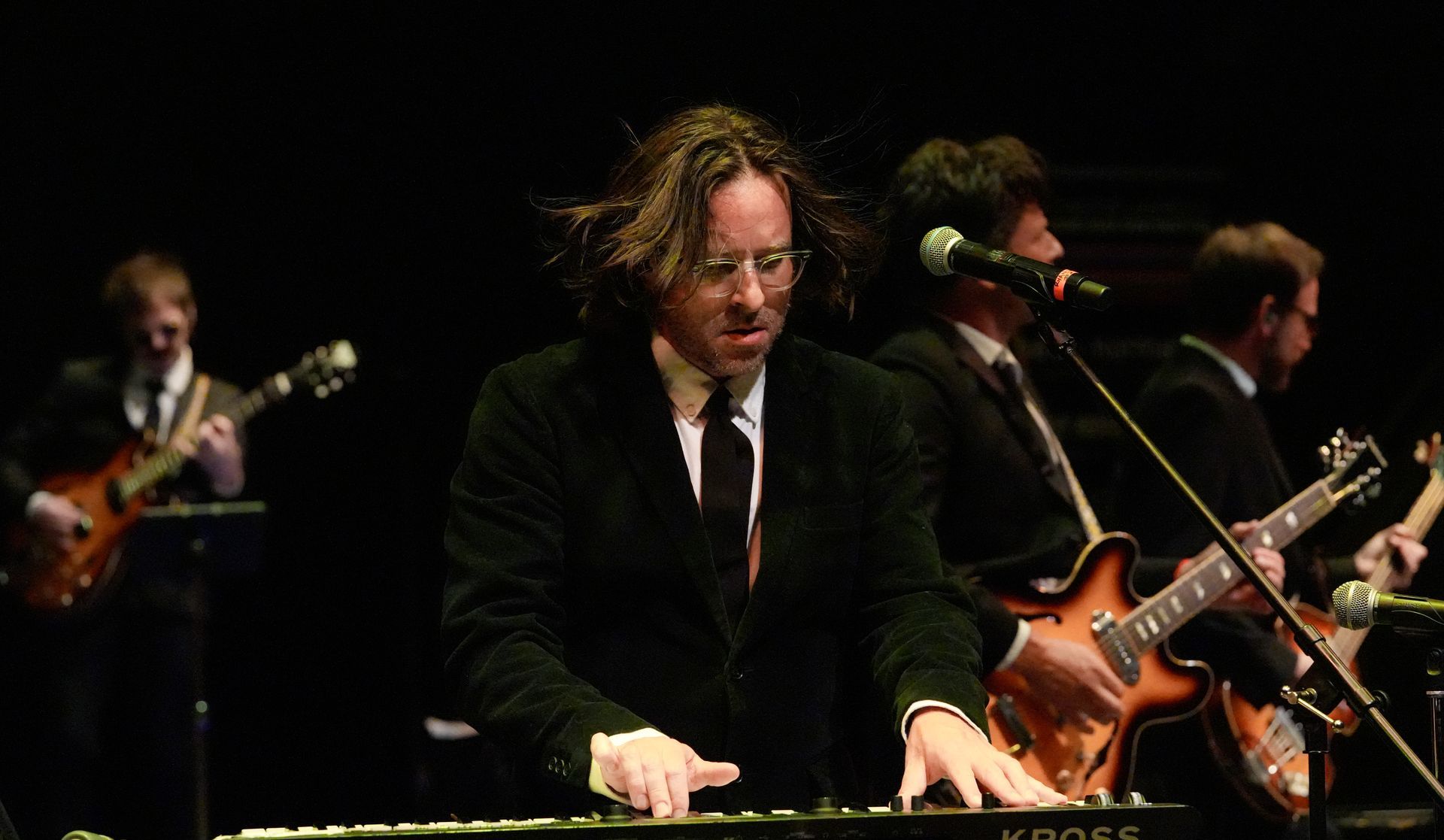 Band performing onstage: keyboardist in focus, electric guitars, dark setting.