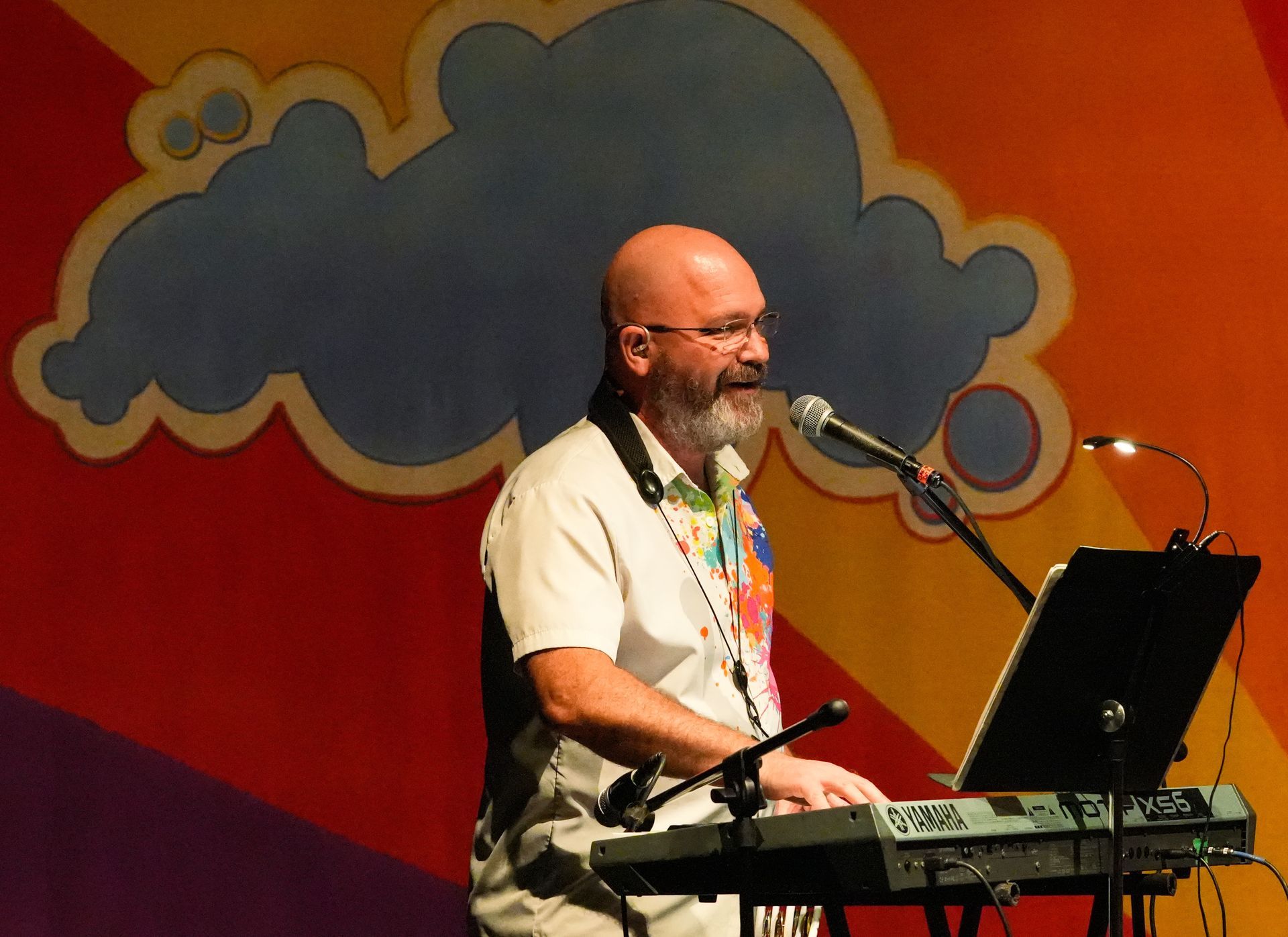 Man with a beard plays keyboard on stage, colorful background with cloud design.