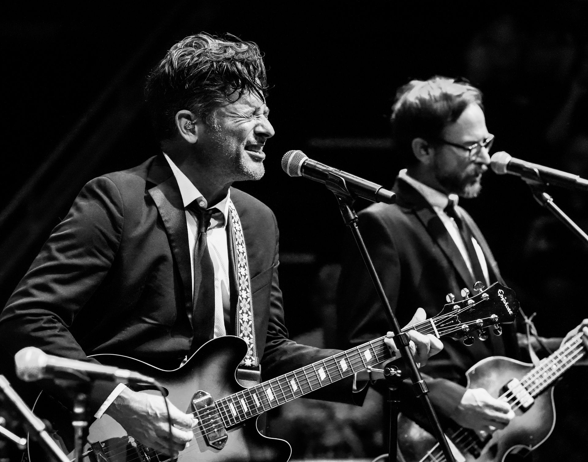 Two men in suits playing guitars on stage, singing into microphones. Black and white.