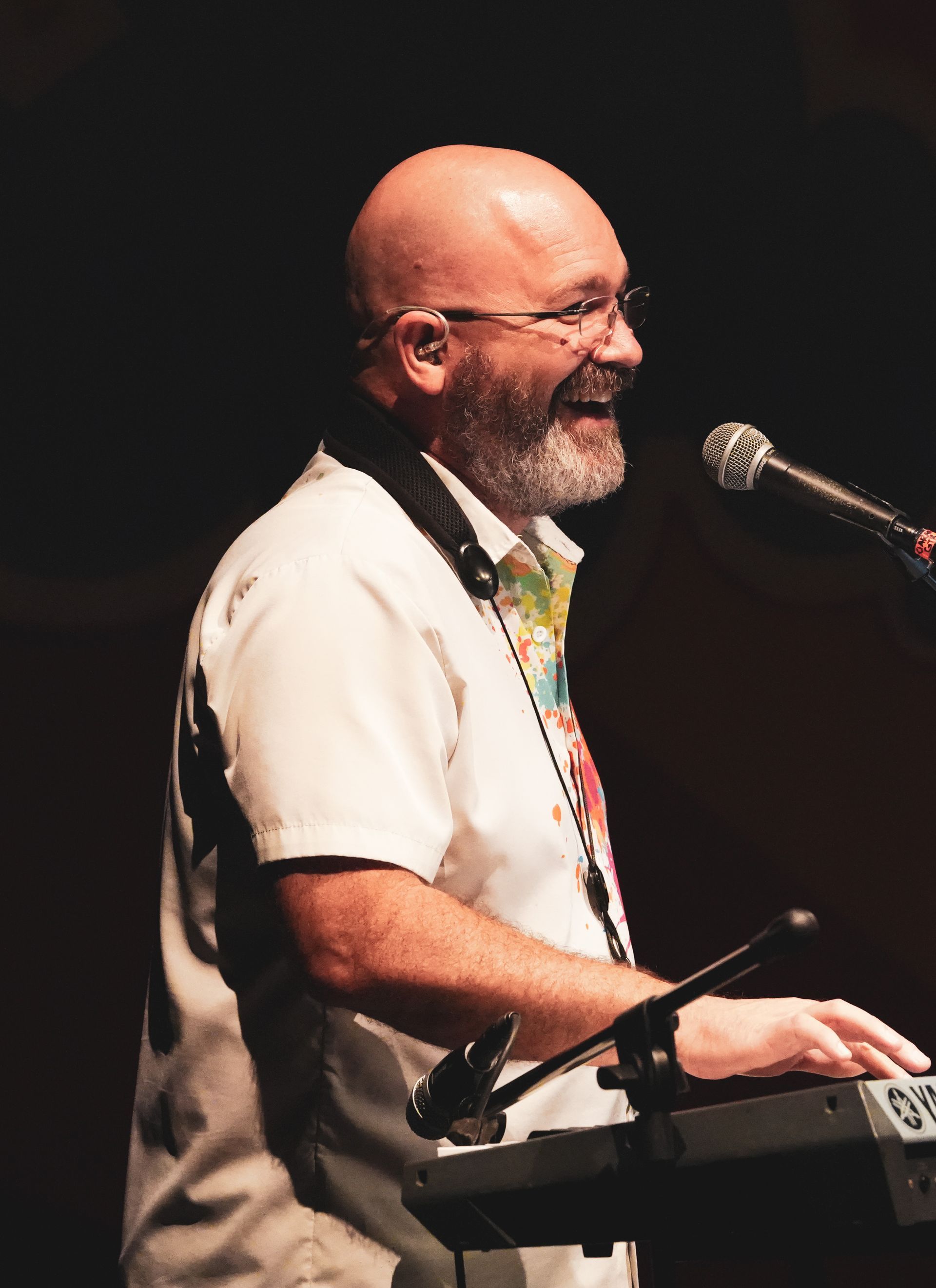 Bald man with glasses plays keyboard, singing into a microphone. Smiling on stage, wearing a white shirt.