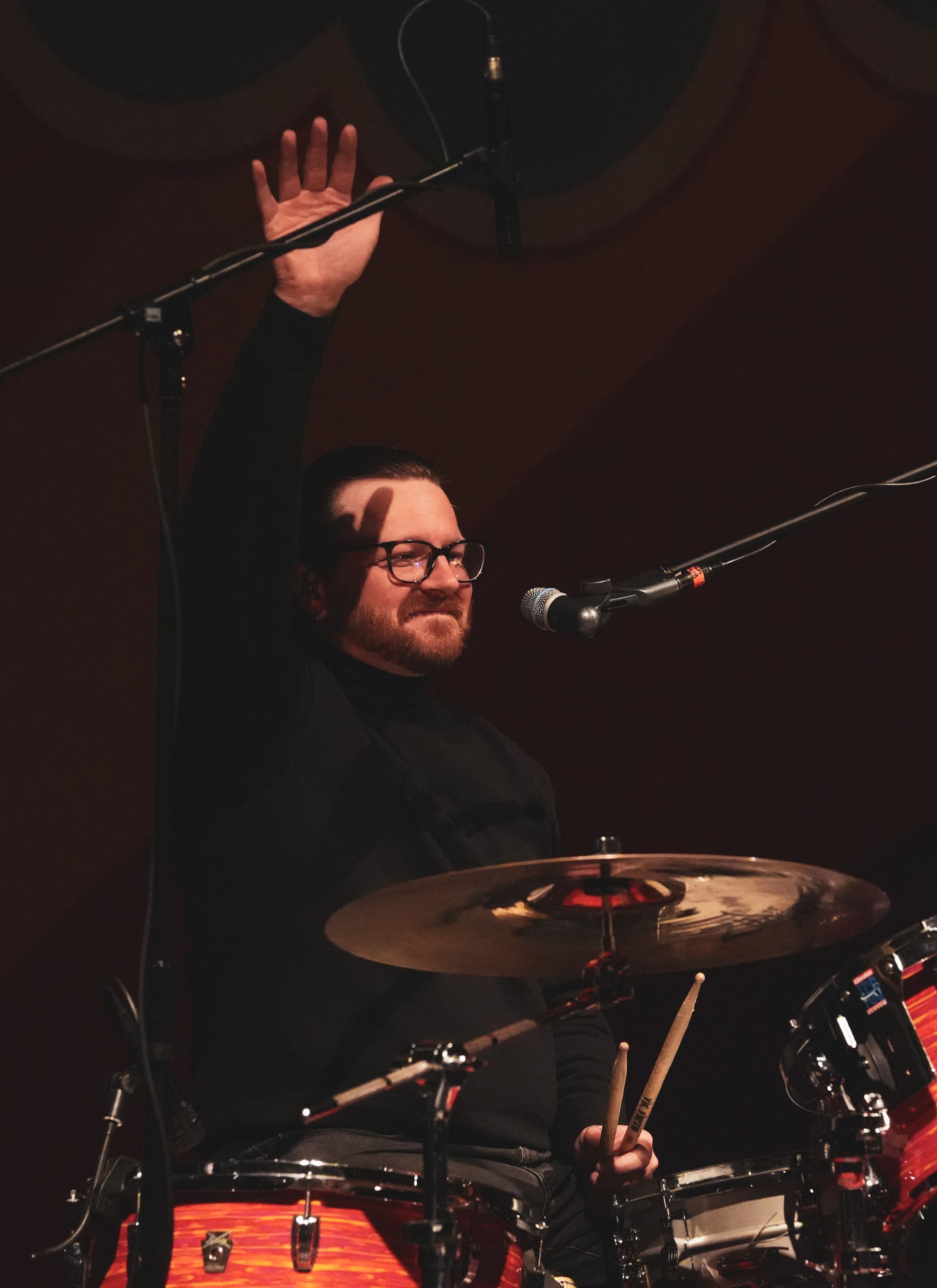 Drummer raising hand, playing drum set on stage. Black clothing, glasses, warm lighting.
