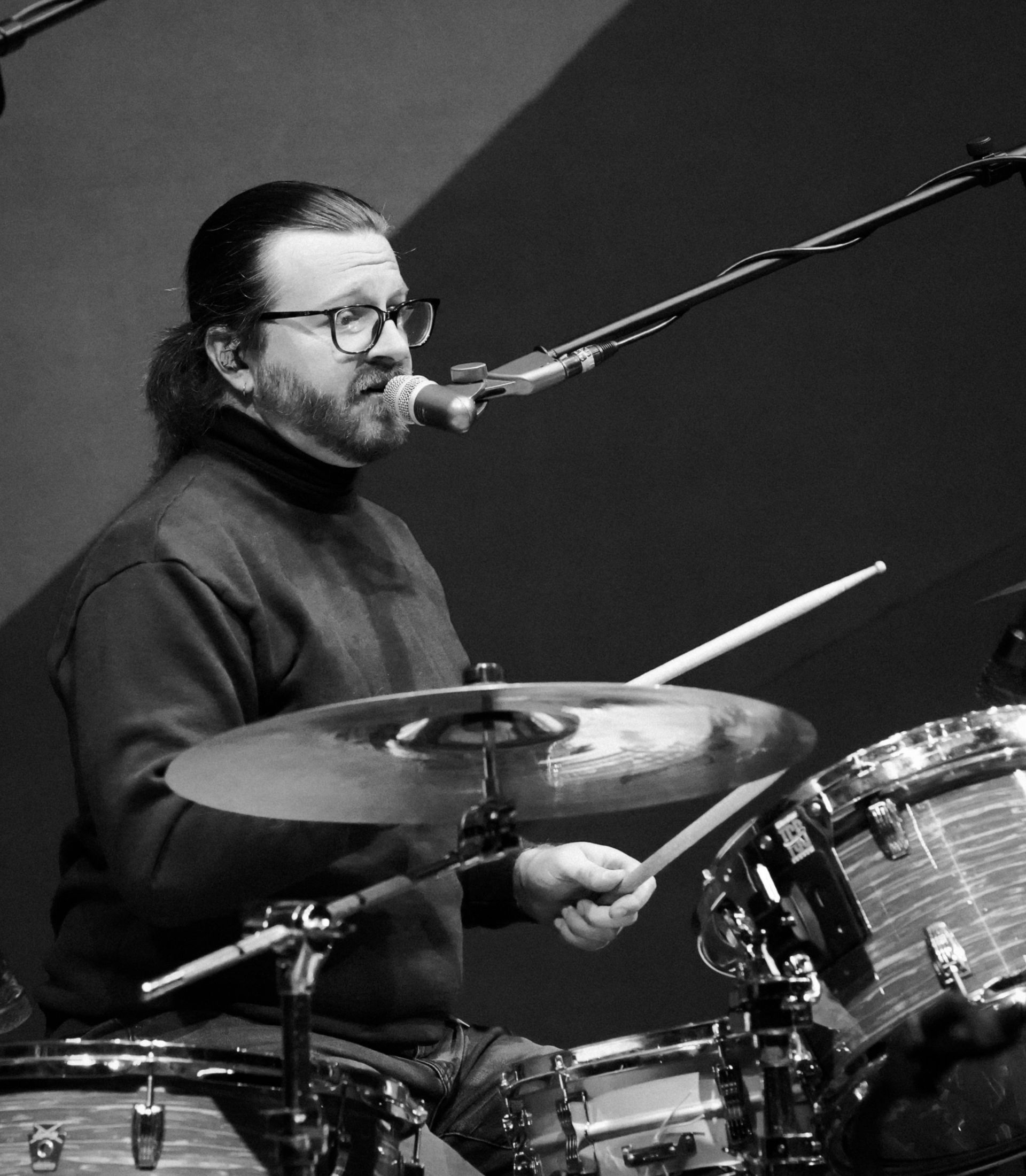 Man playing drums, wearing glasses and a turtleneck. Performing in a dim setting, holding drumsticks, and near a microphone.