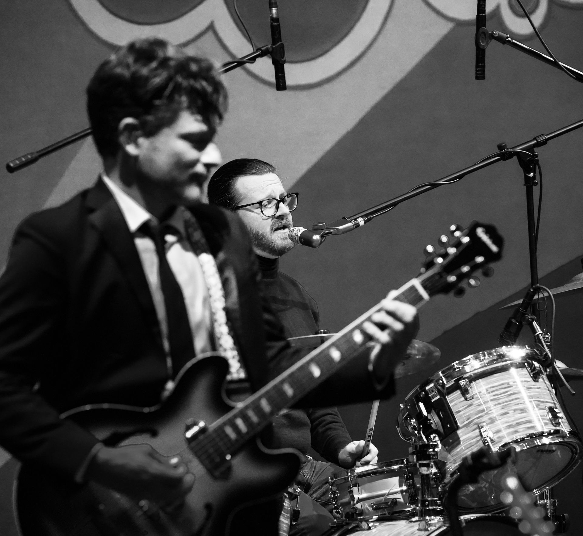 A band performs live, guitarist in focus, singer behind a microphone, drum set visible.