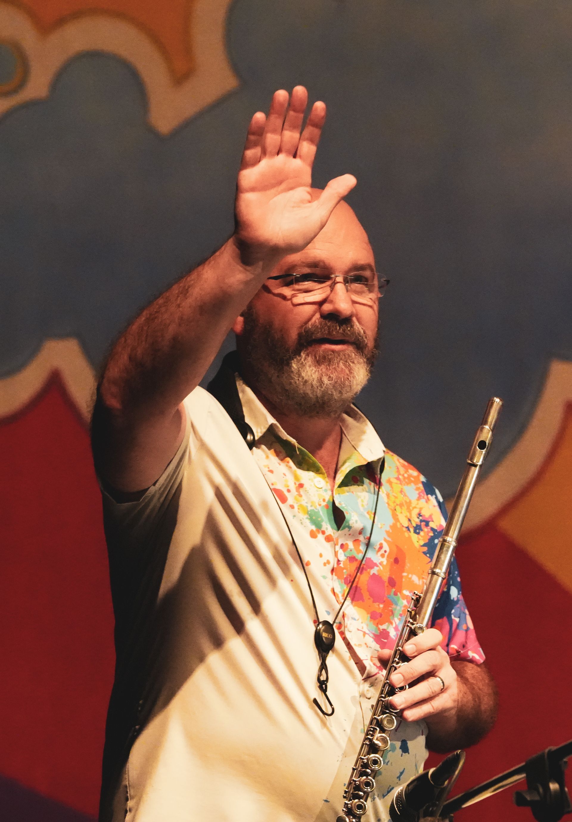 Man with beard waves, holding a flute. He wears a colorful shirt, stands on stage.