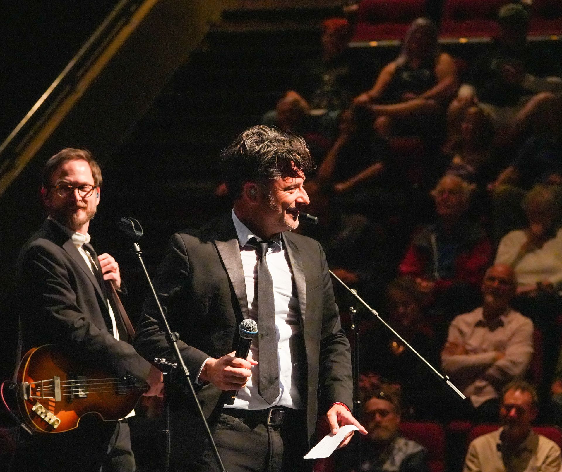 Two men in suits on stage, one with a guitar, speaking to an audience.