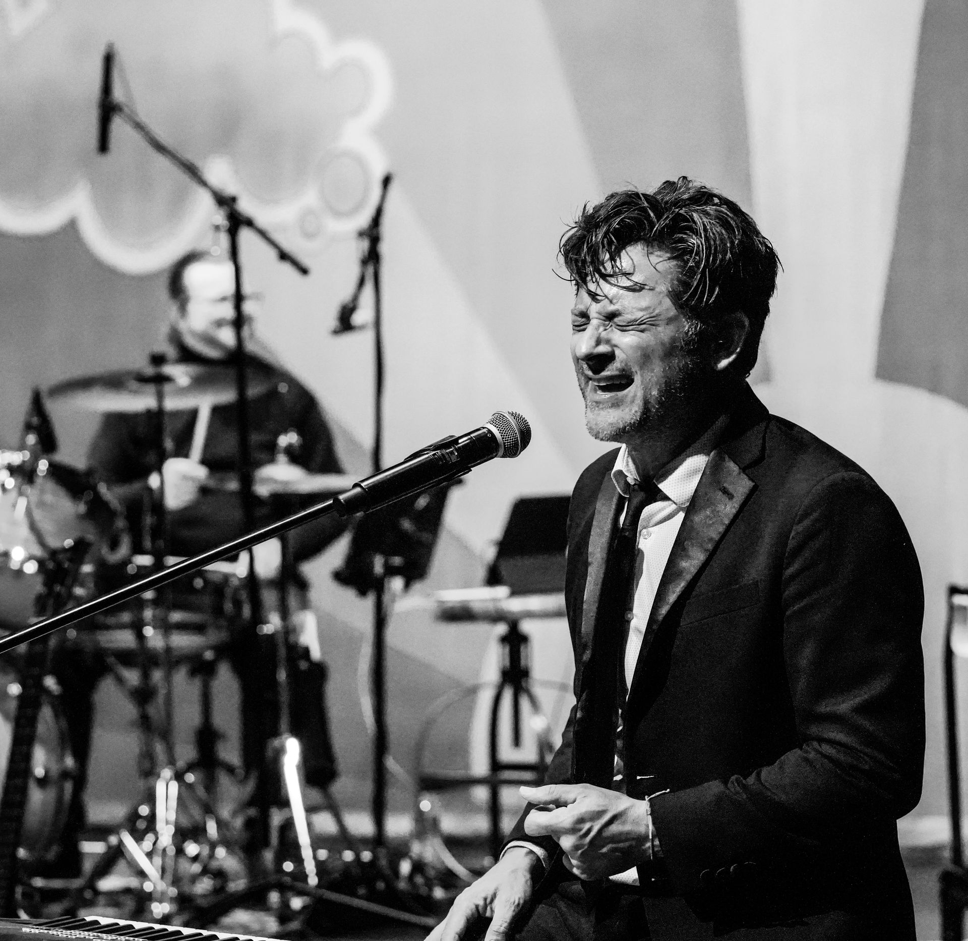 Man singing into a microphone onstage; drummer in background. Black and white.