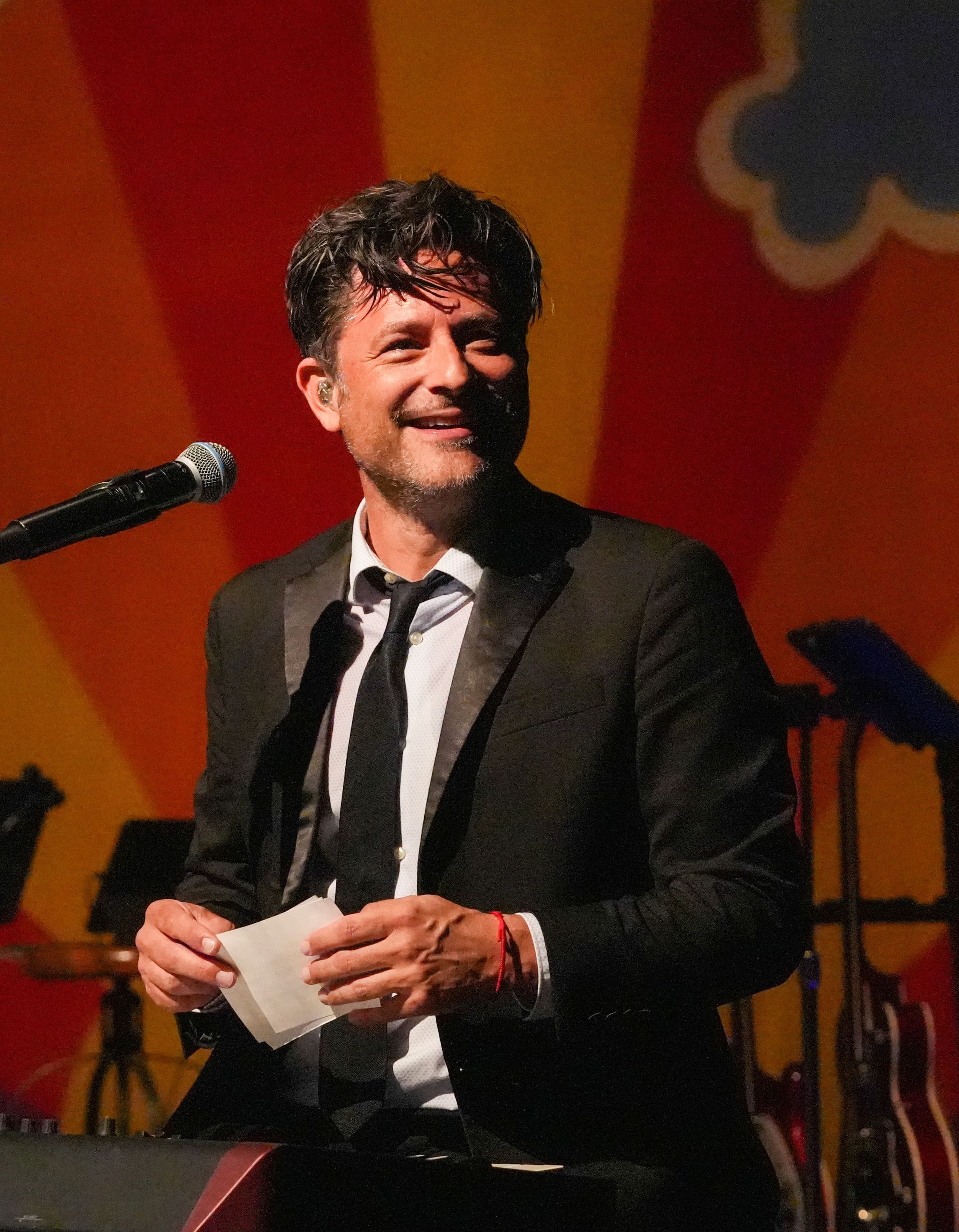 Man in suit on stage, holding paper. Red and yellow background, instruments visible.