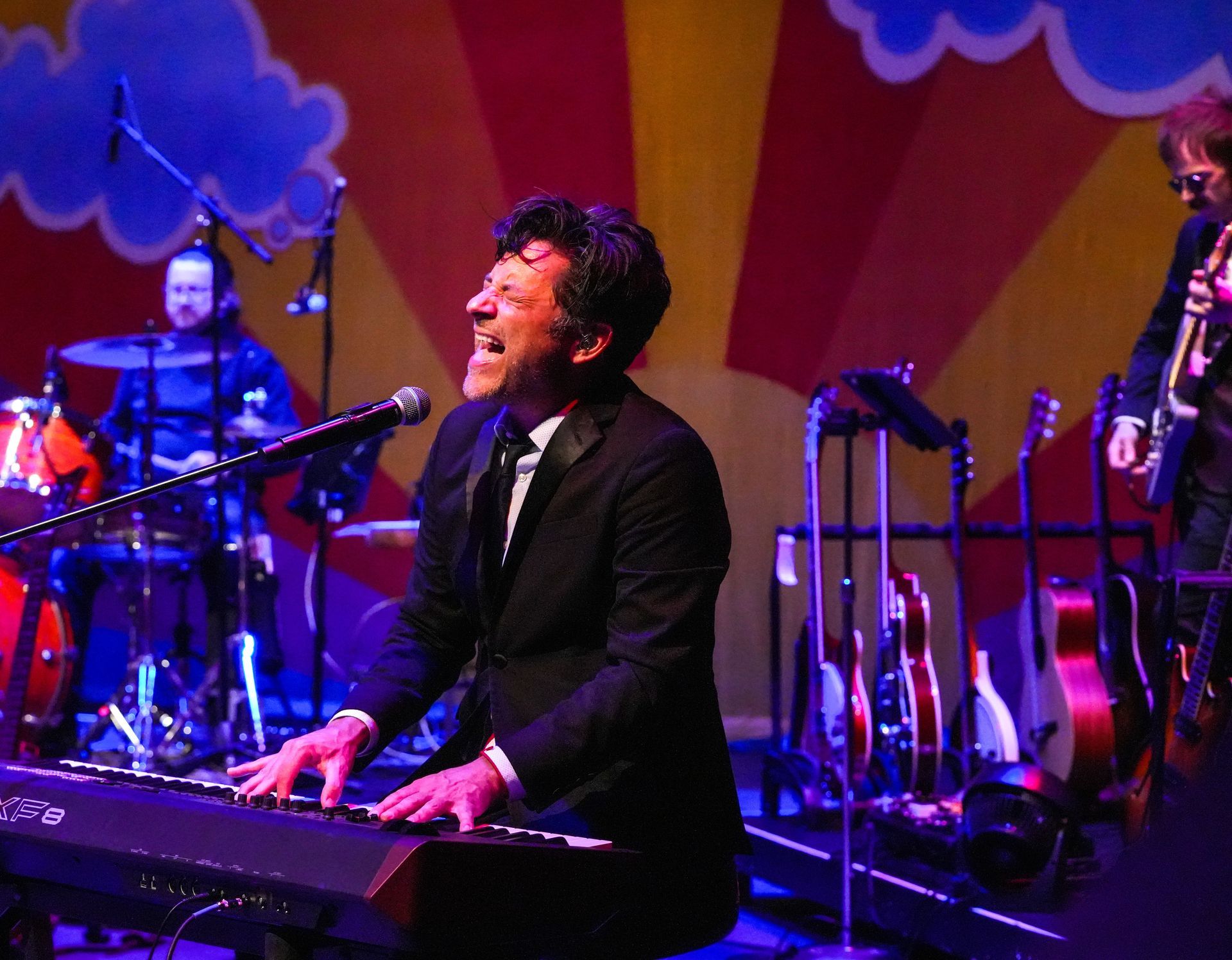 A musician in a suit plays a keyboard on stage, singing. Colorful backdrop and band members visible.