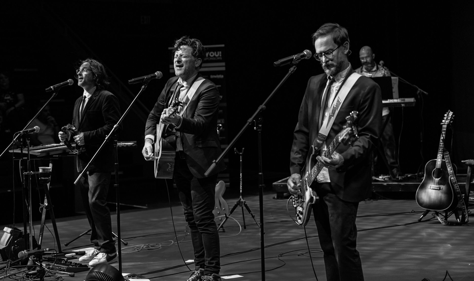 Band on stage, performing with guitars and microphones in a dim setting. Black and white photo.