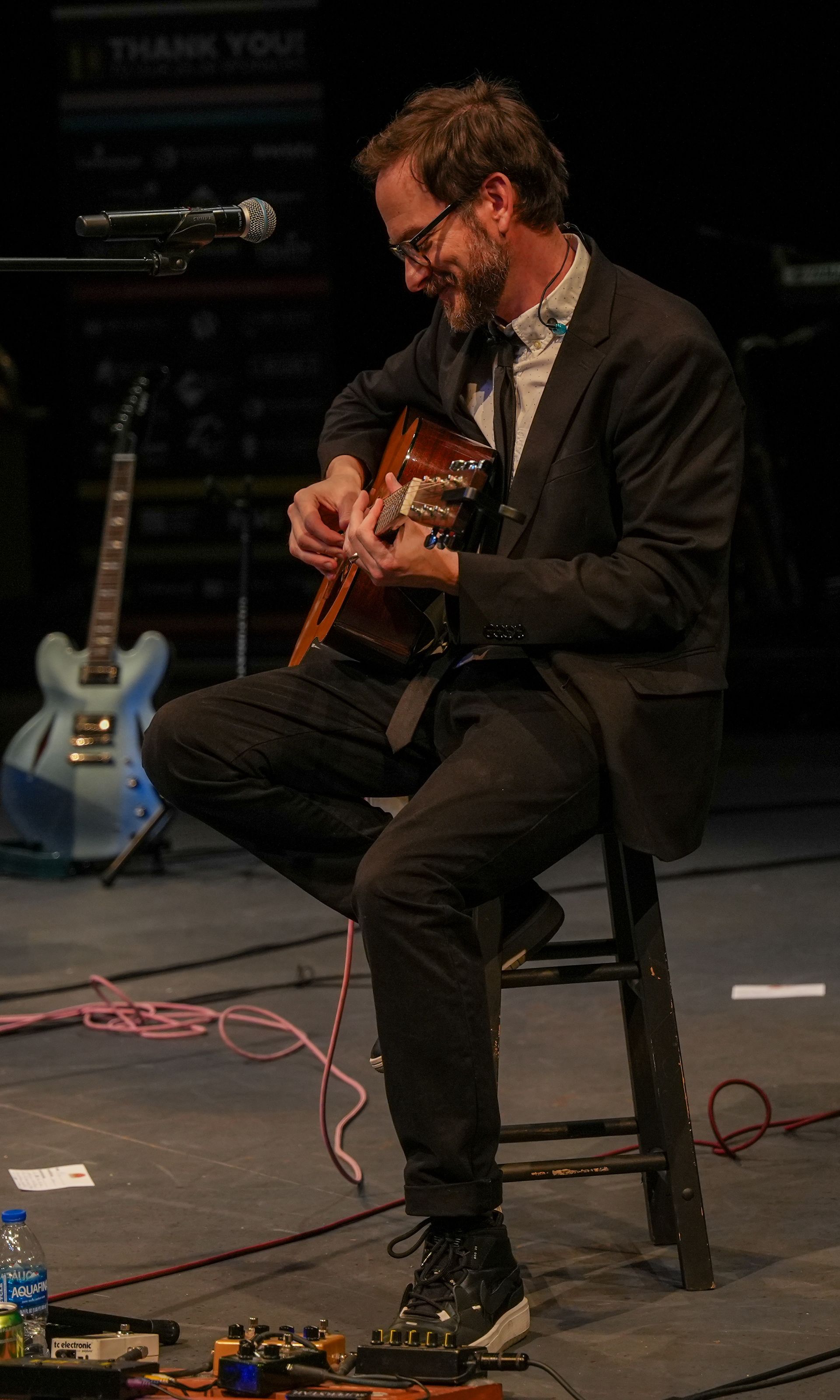 Man in suit playing guitar, smiling. Seated on a stool onstage, electric guitar in background.