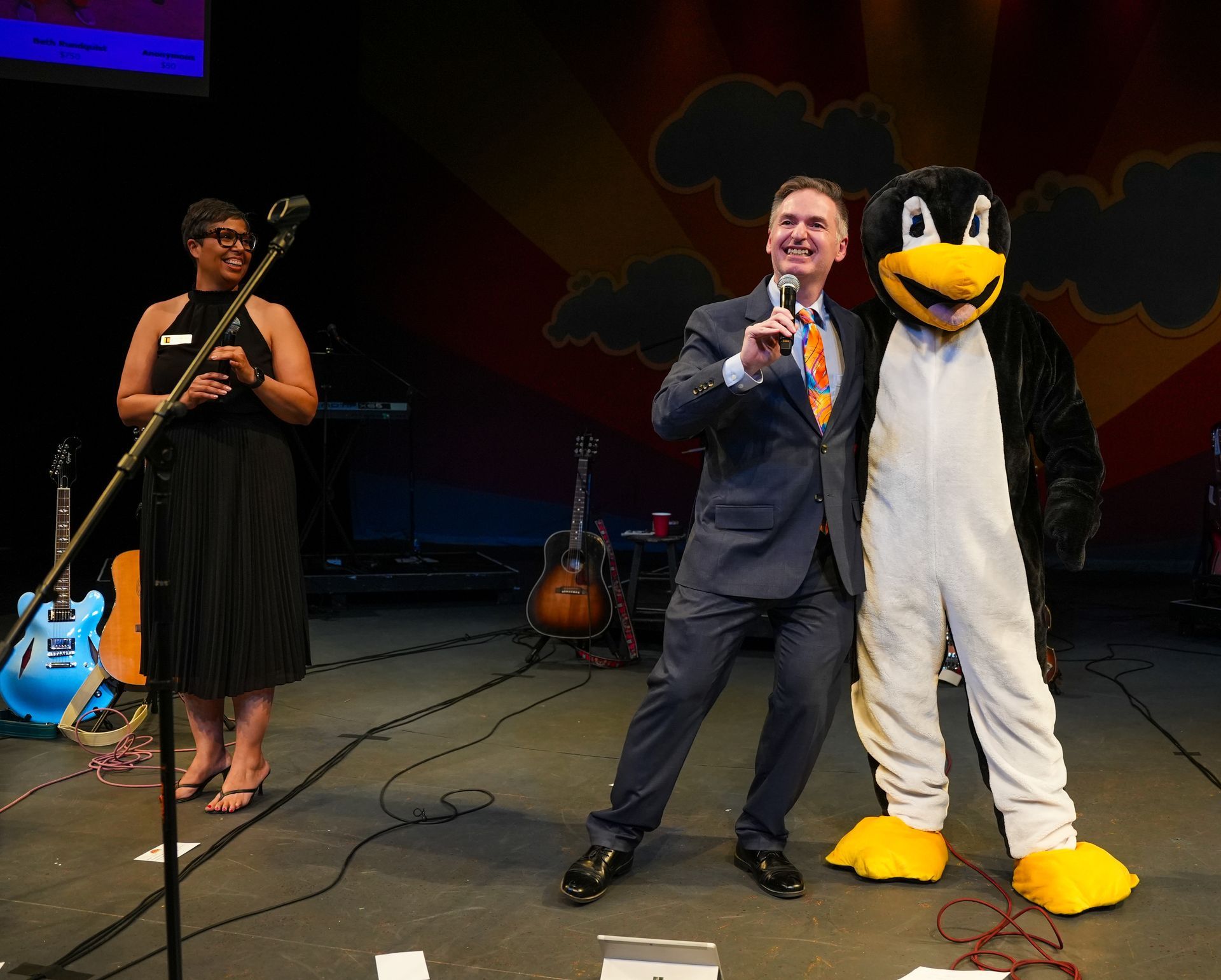 Man in suit smiles, poses with penguin mascot on stage; woman stands nearby.