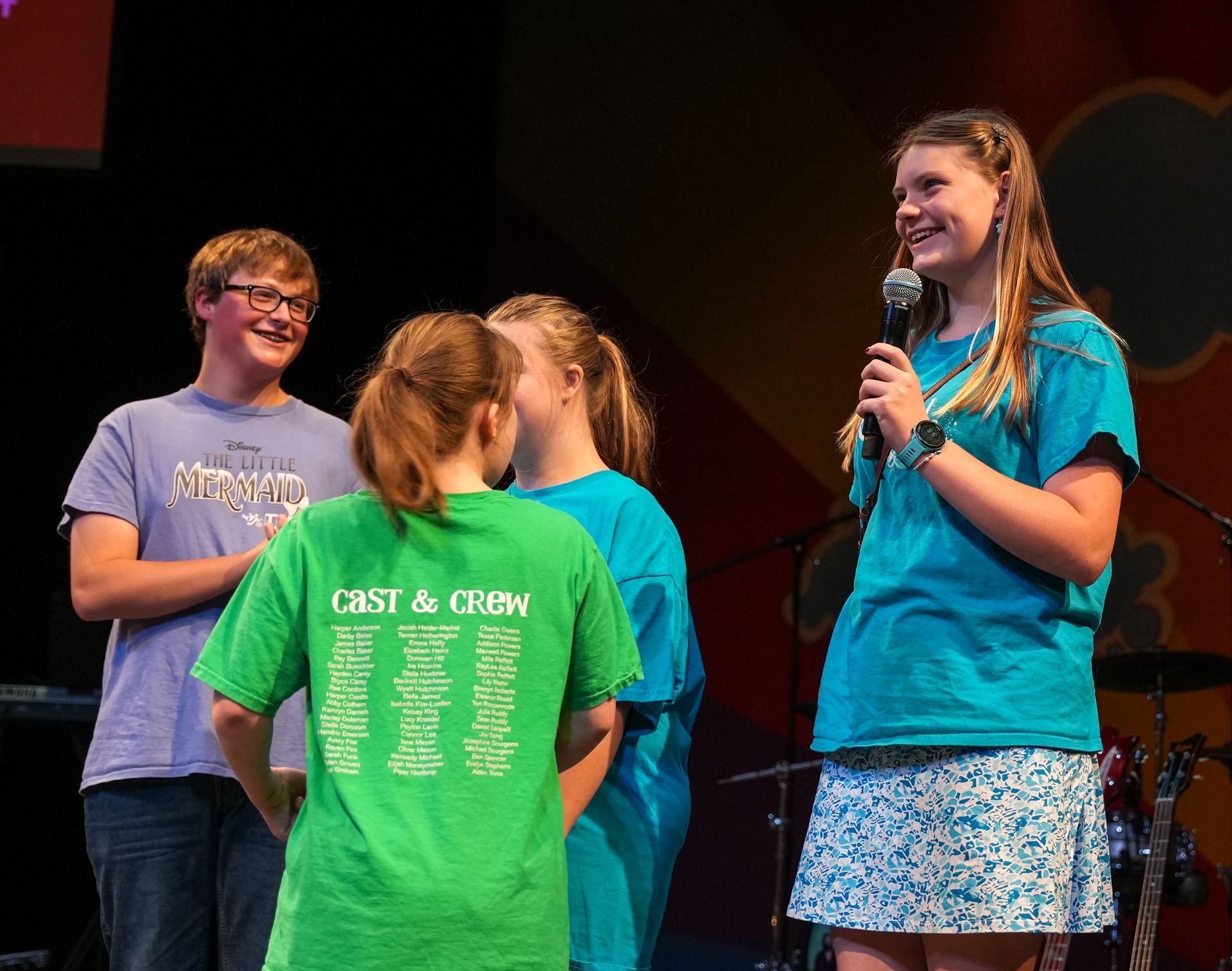 Four young people onstage; one with microphone smiles, others listen. Teal and green shirts.