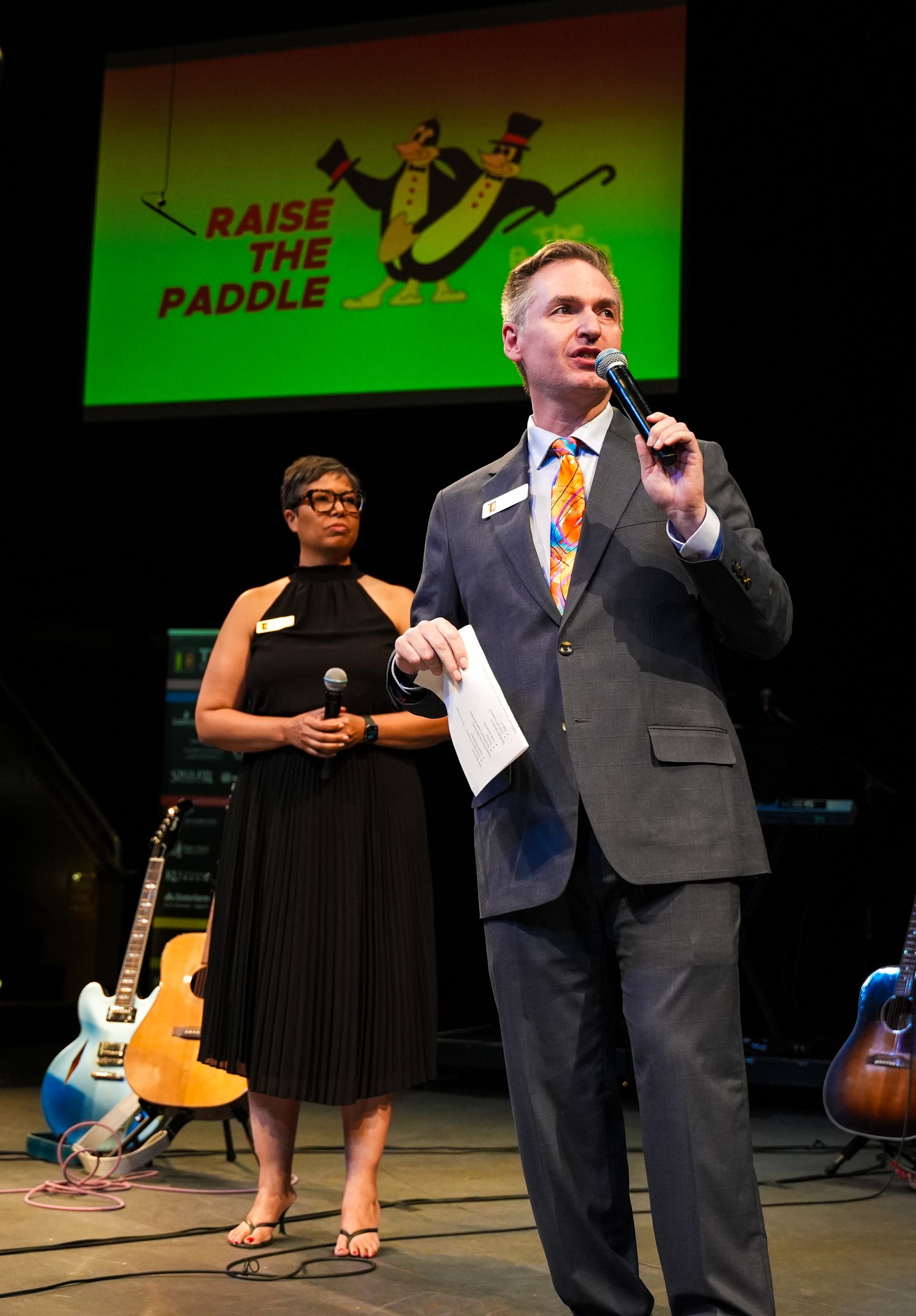 Man speaks into a microphone at an event with a woman standing beside him.