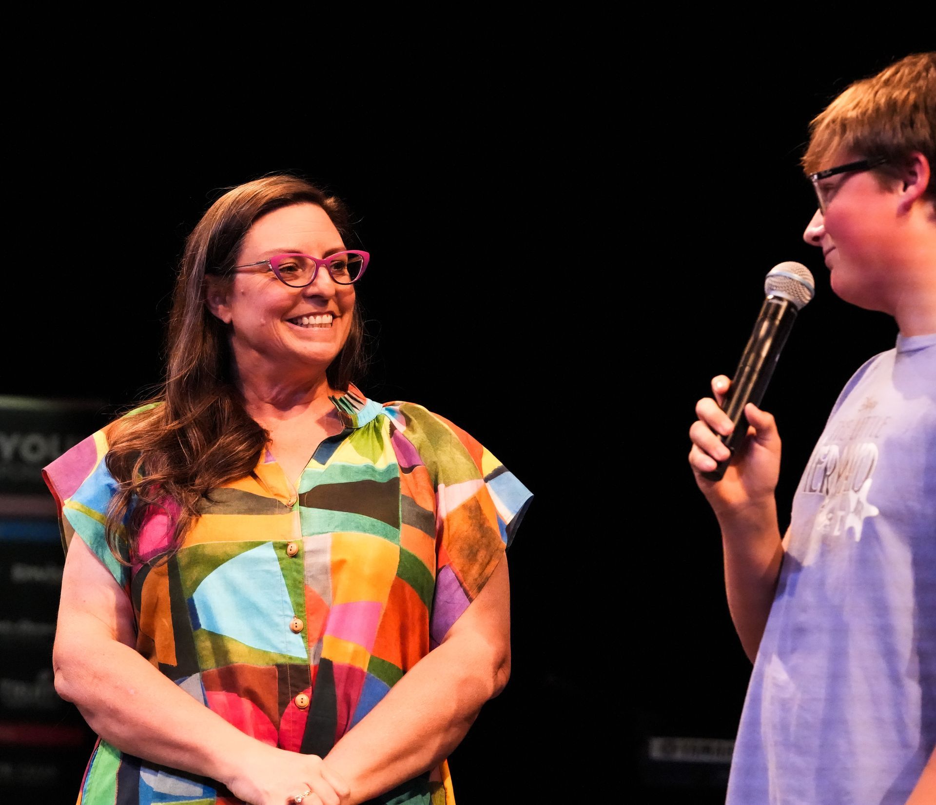 Woman in colorful shirt smiles at a boy holding a microphone on a stage.