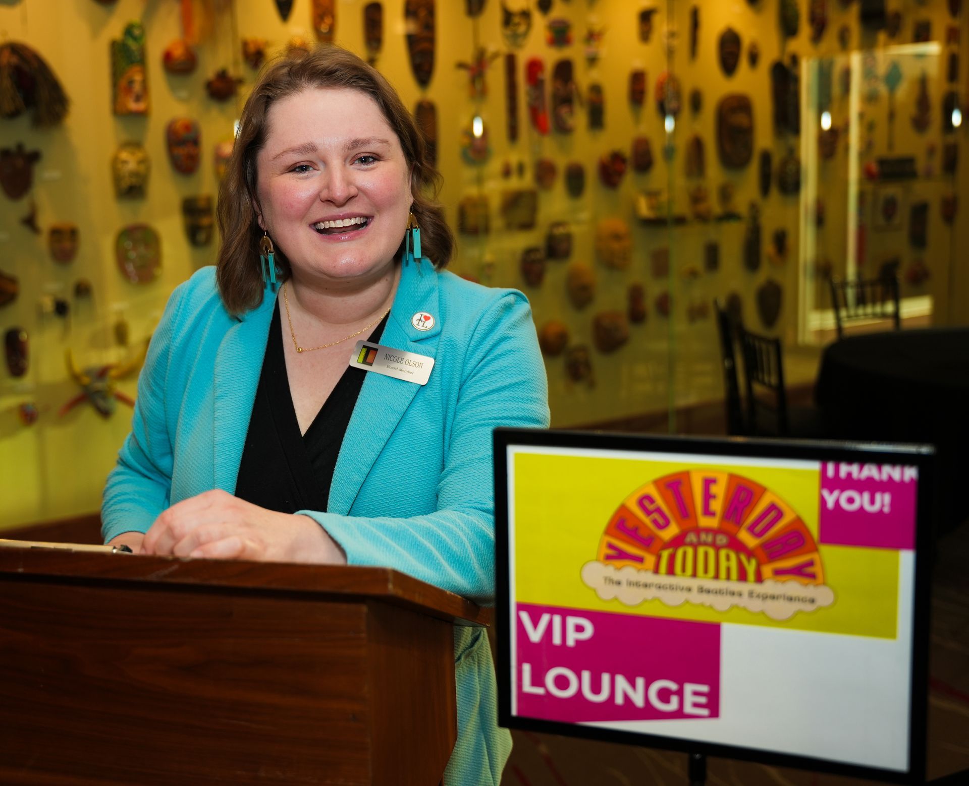 Woman in turquoise blazer smiling, standing at a VIP Lounge sign in front of a wall with masks.