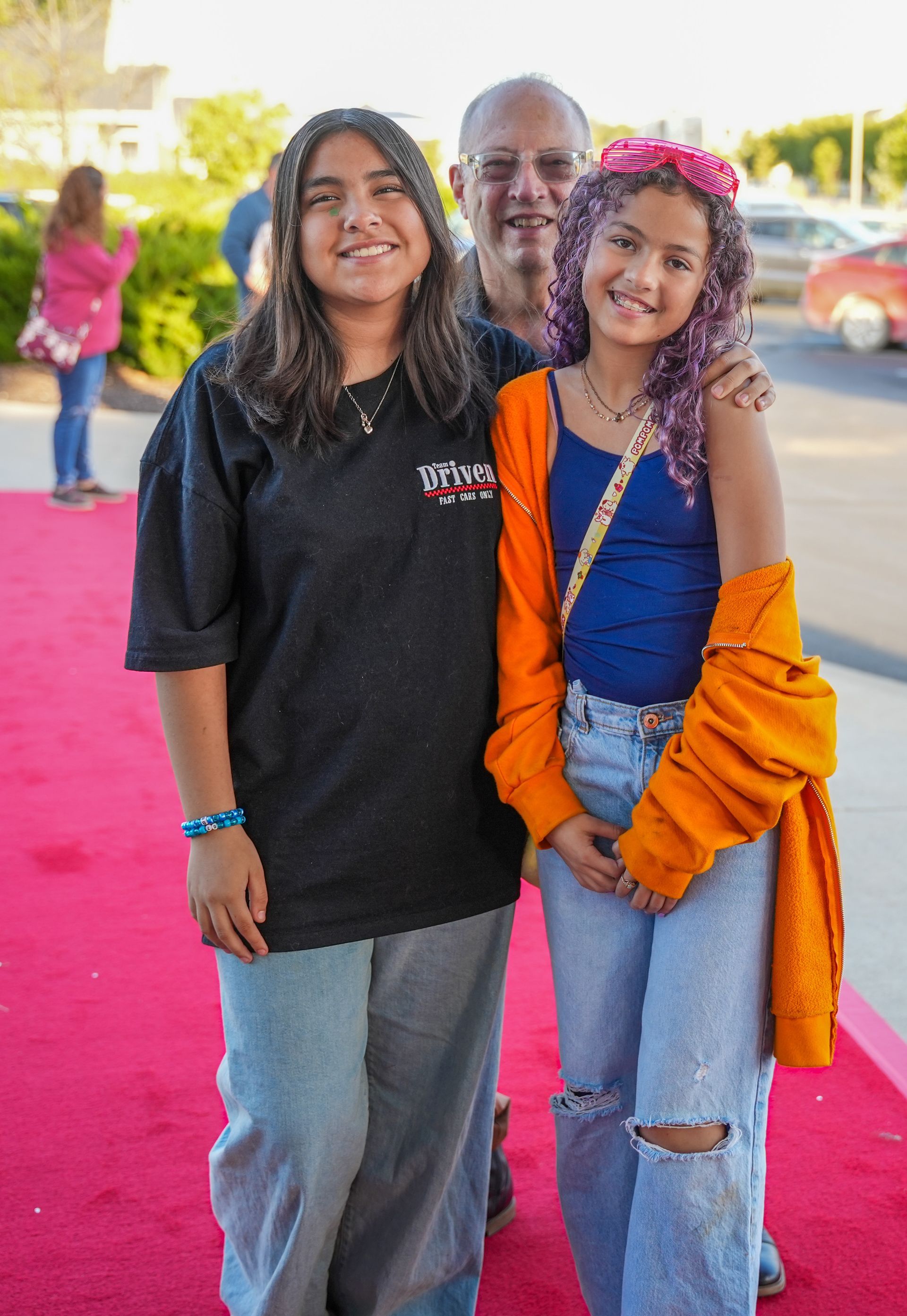 Two smiling girls and a man on a pink carpet; the girls are wearing casual outfits.
