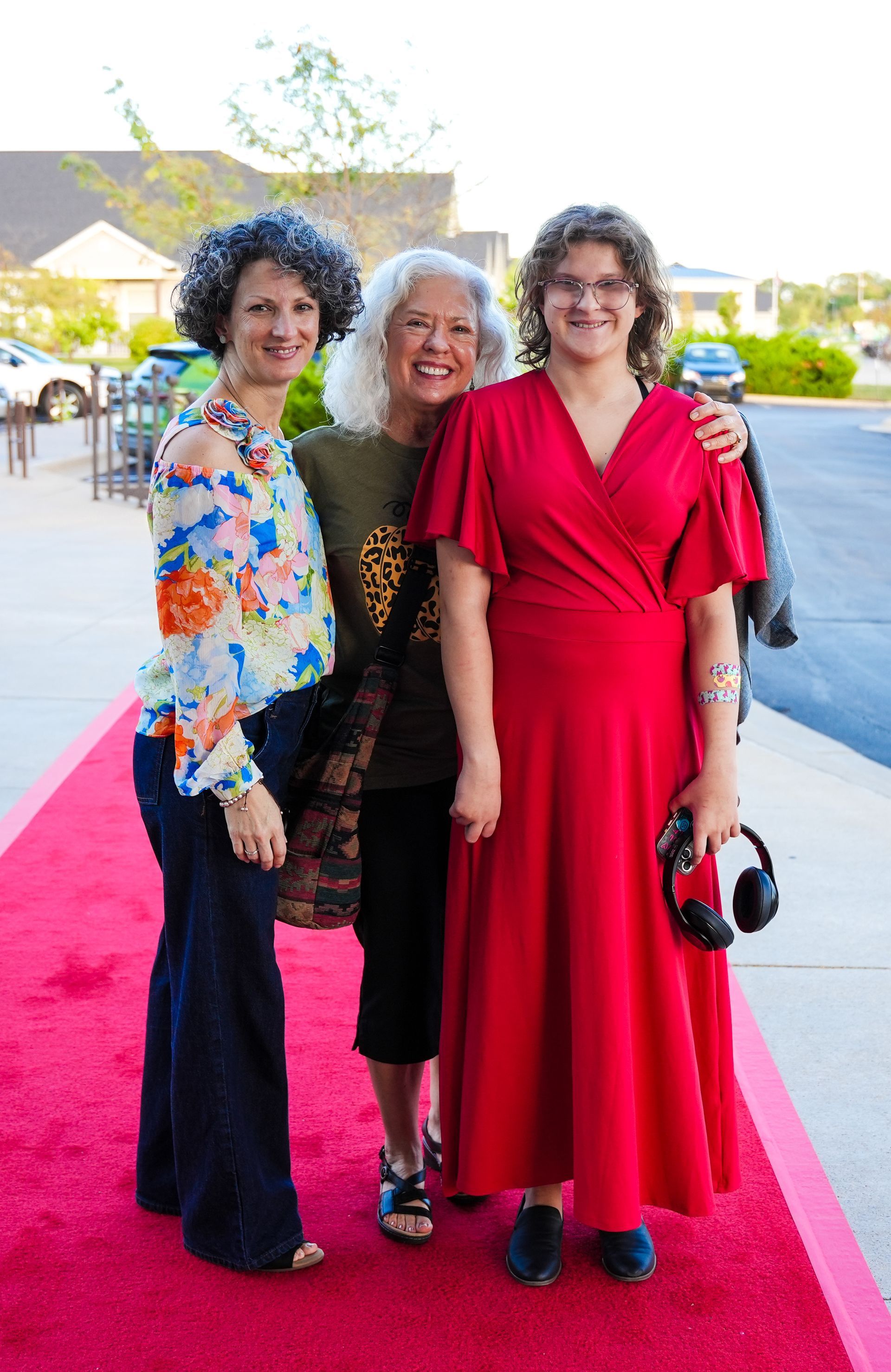 Three people standing on a red carpet smiling. A woman in a red dress holds headphones.
