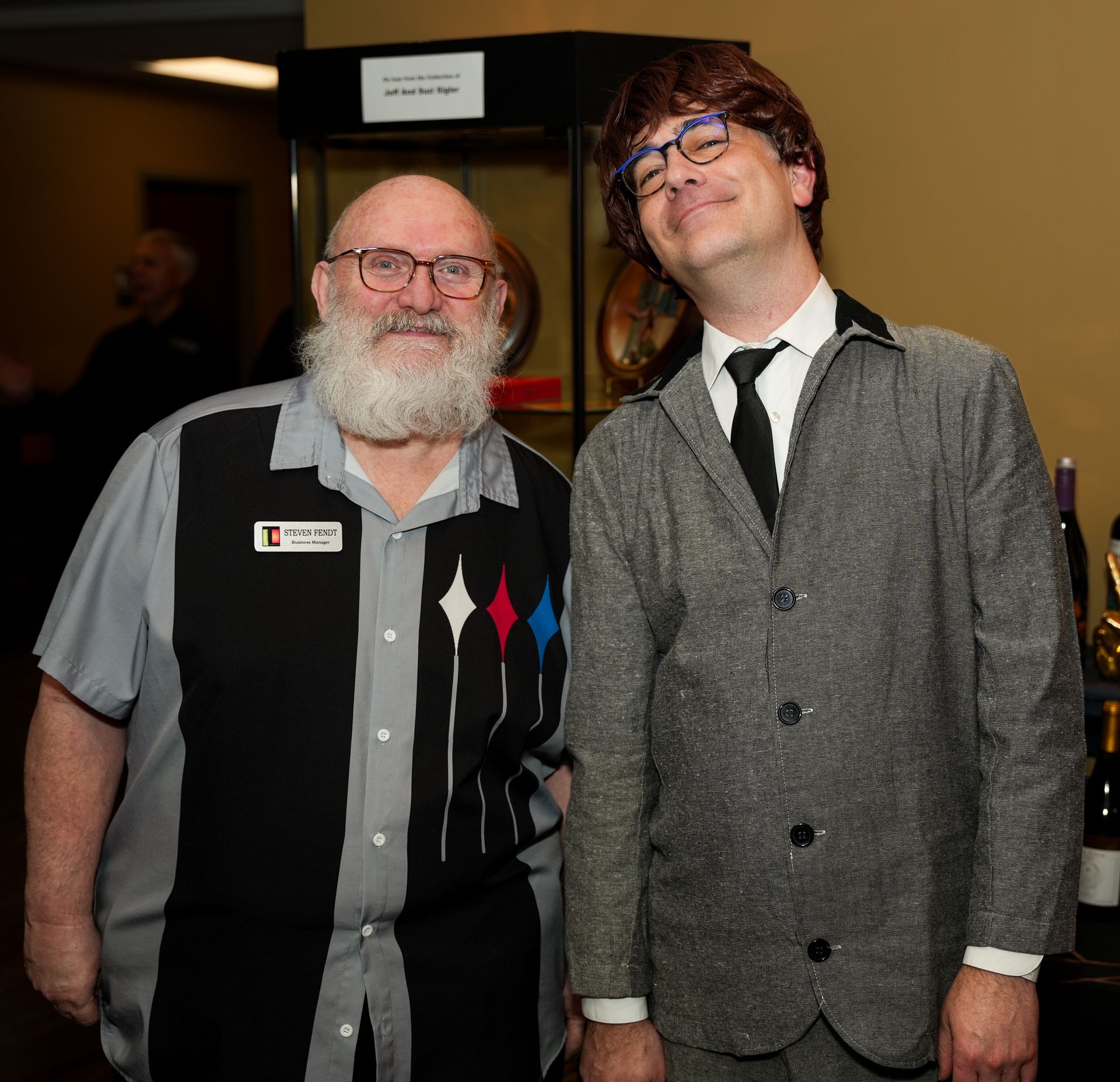 Two men posing. One with a white beard wearing a bowling shirt, the other in a gray suit with a wig.