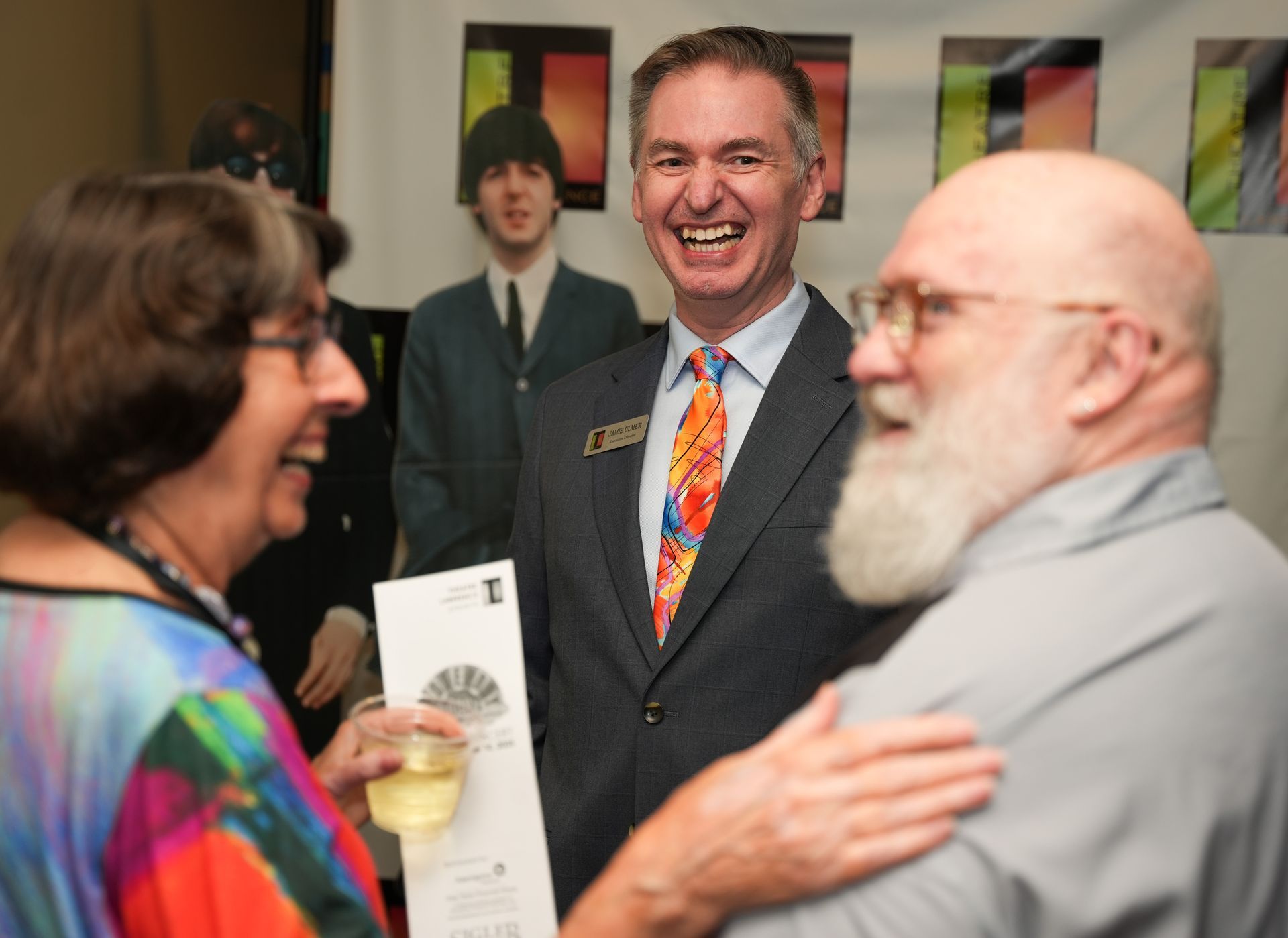 Man in suit laughs with two people at an event; Beatles poster in background.