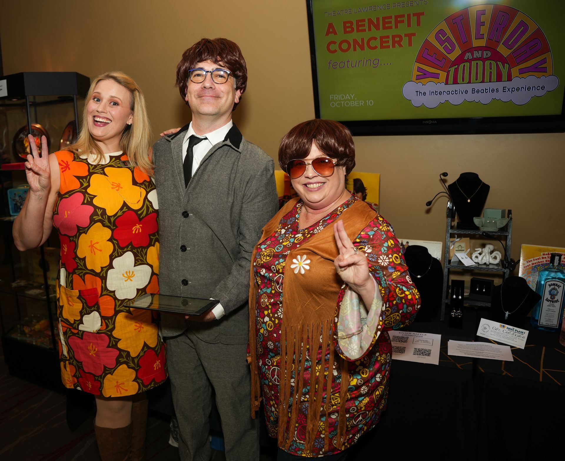 Three people in 60s costumes, posing for a photo at a benefit concert, all giving peace signs.