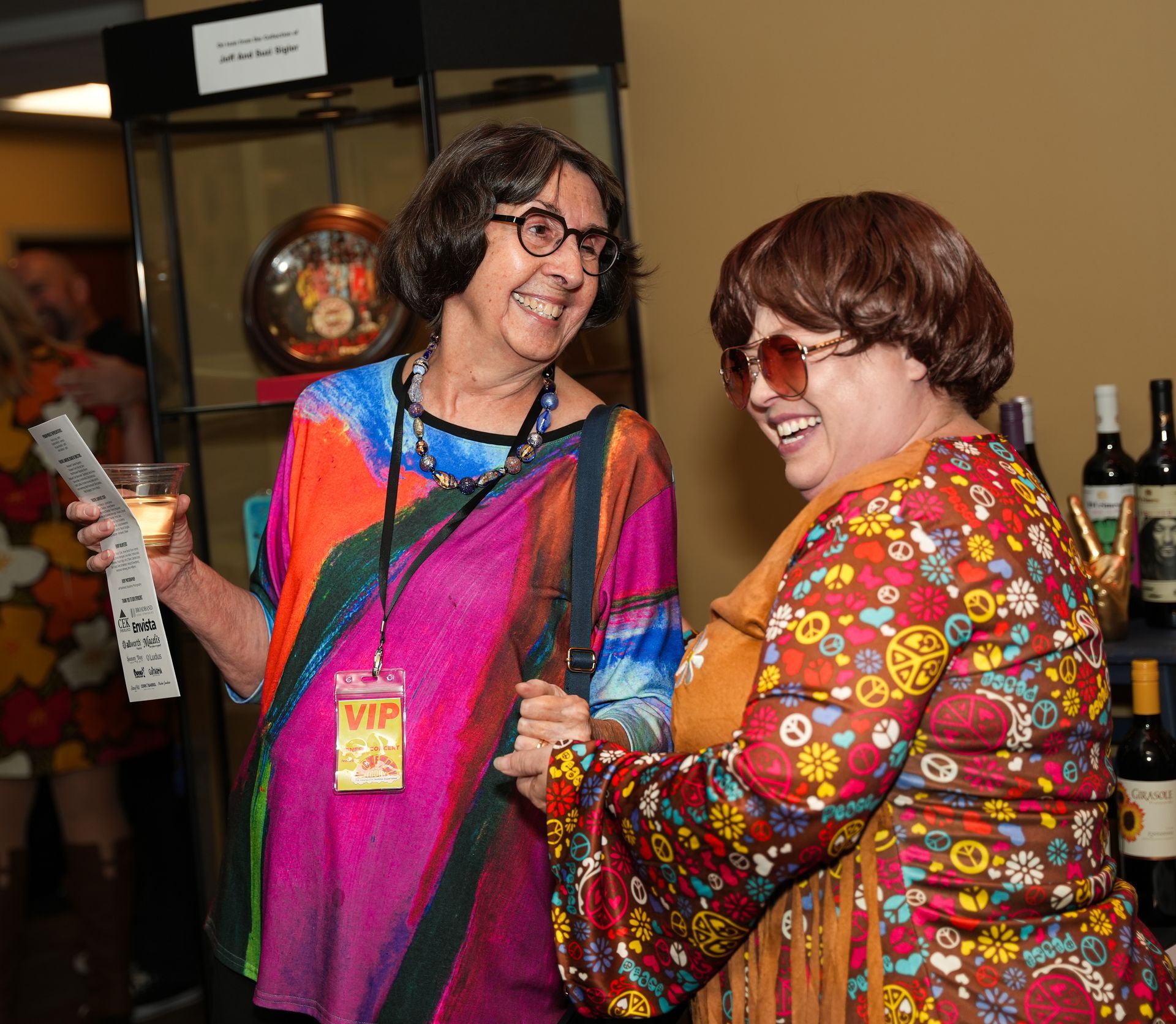 Two people in colorful 60s costumes laughing, holding drinks. One wears a VIP pass.