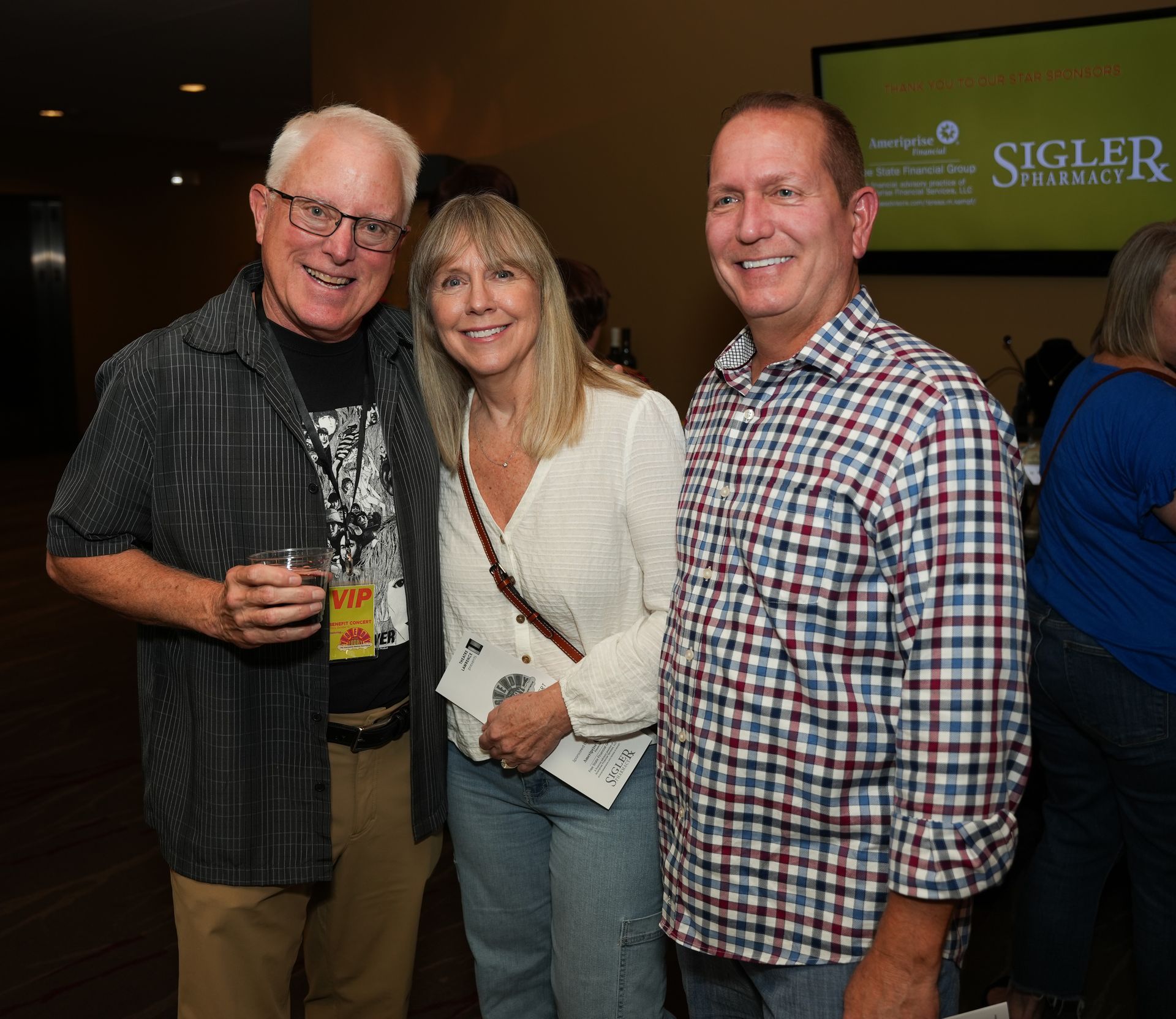 Three people smiling, posing together indoors. A man on the left holds a drink, the woman in the middle holds a pamphlet.