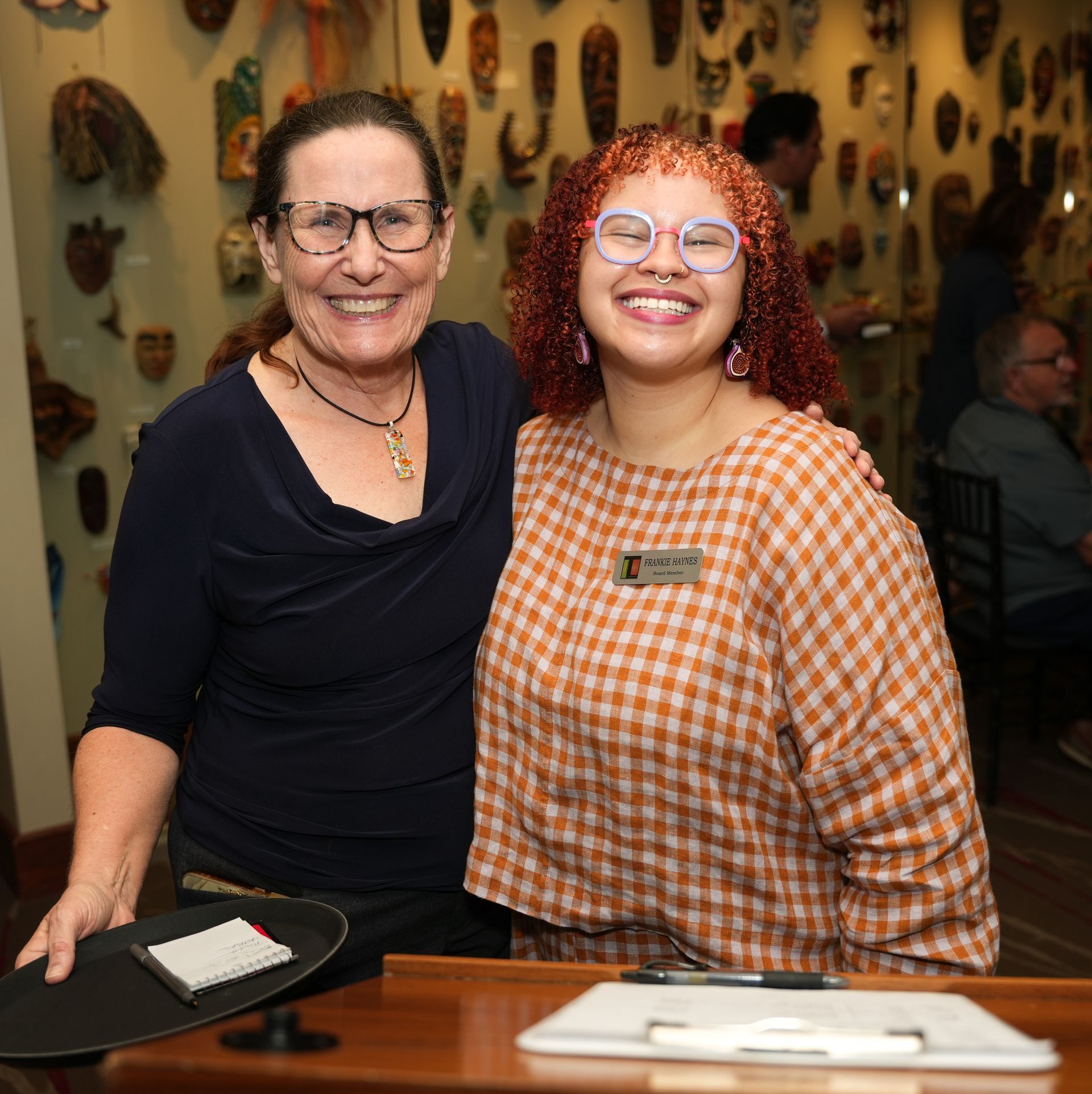 Two women smiling at a desk. One has dark hair and glasses; the other has curly orange hair and glasses, wearing a name tag.