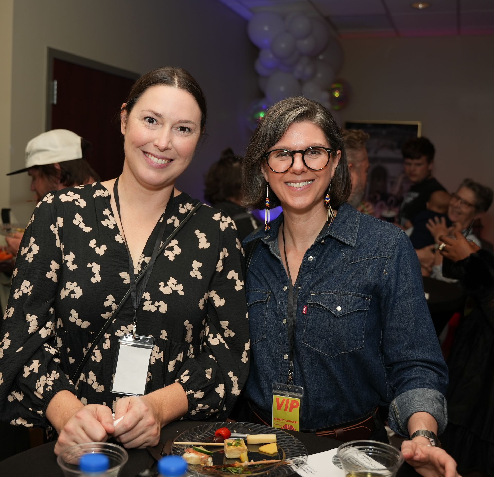 Two women smiling at a table, one in a floral top, the other in a denim shirt, with food and drinks in an event setting.