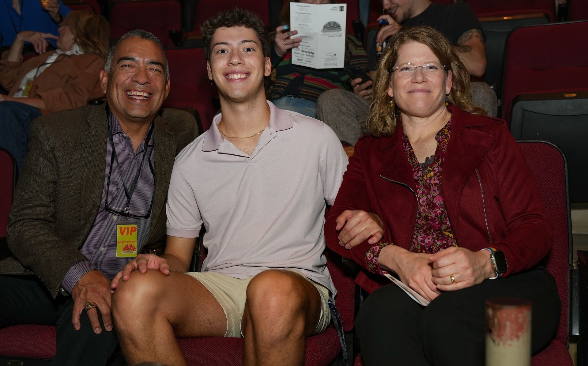 Three people seated in theater: Man with blazer, young person in polo, and woman with red jacket, all smiling.