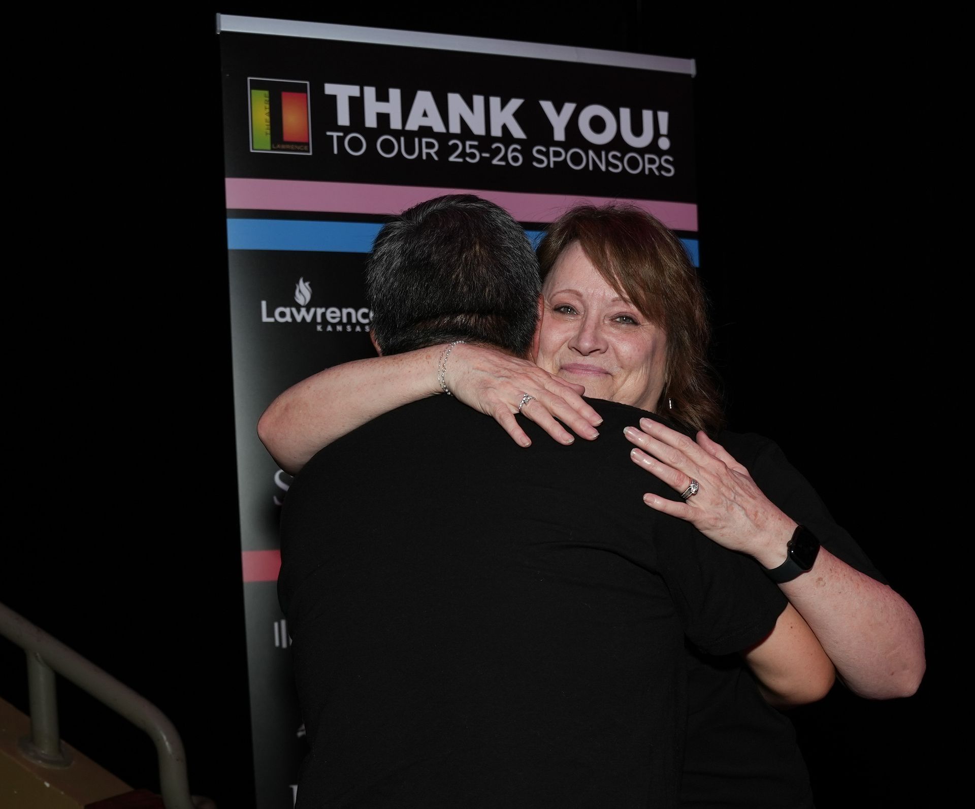 Woman hugs a person with their back to the camera, in front of a banner that says