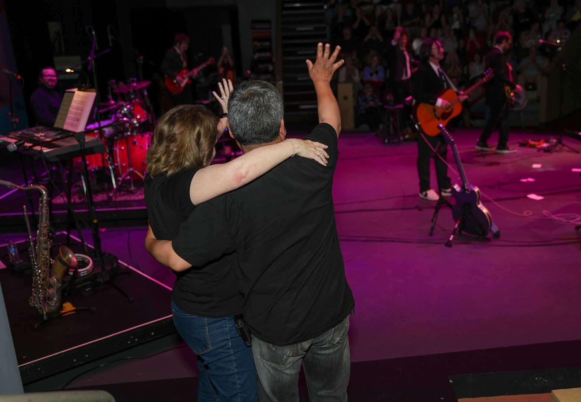 Couple with arms raised facing a band onstage; purple and pink stage lights.