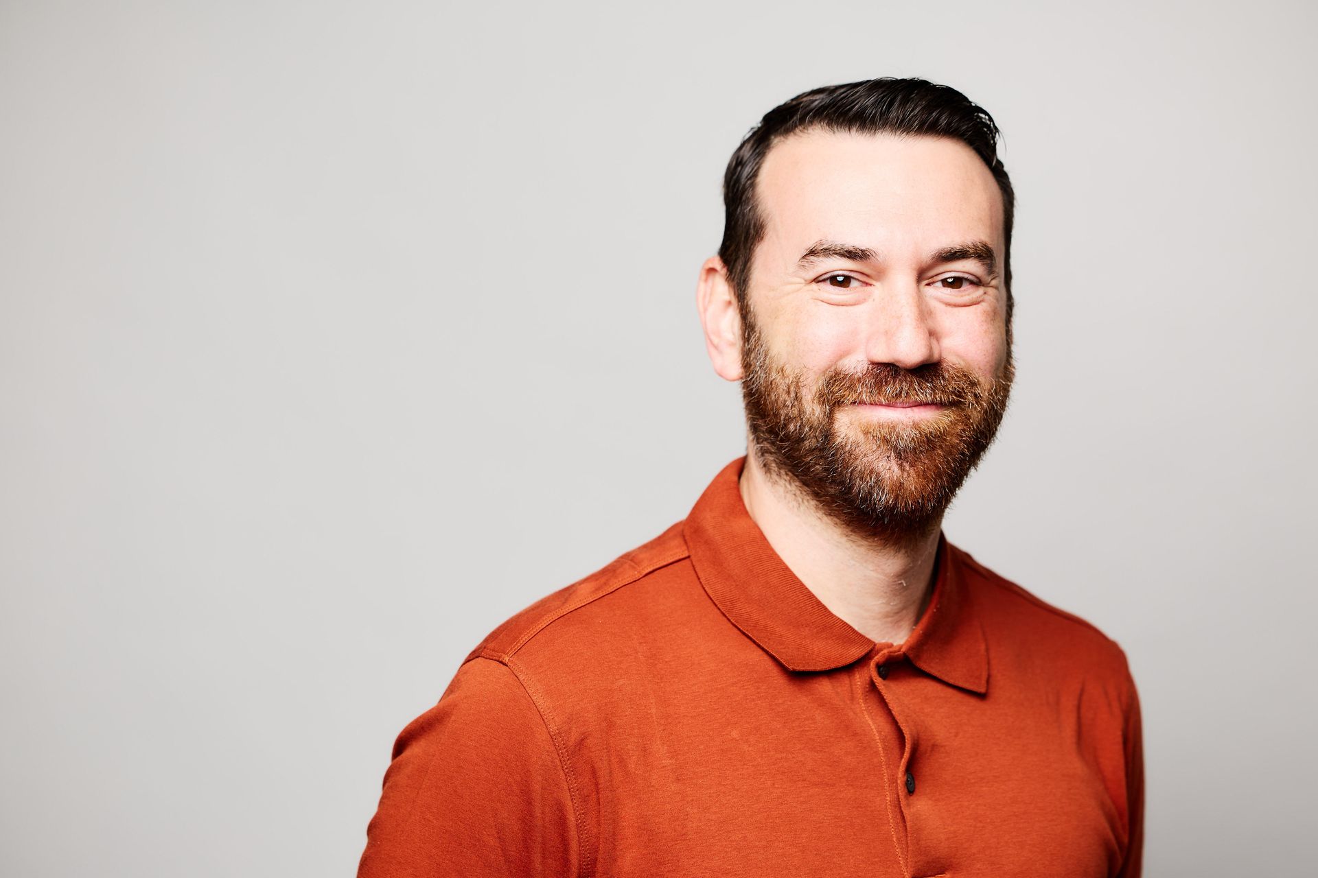 A man with a beard is wearing a red polo shirt and smiling.