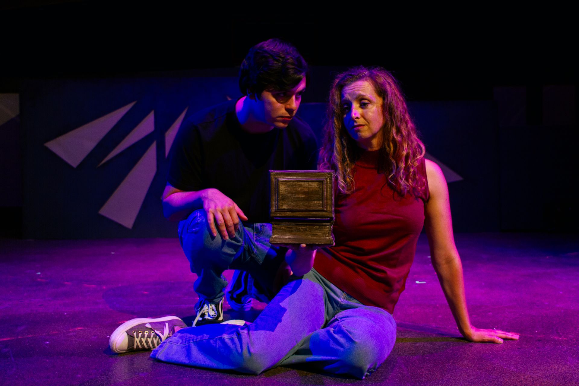 Two people on stage, looking at a small box. Purple stage lighting. Man kneels, woman sits, both in casual clothes.