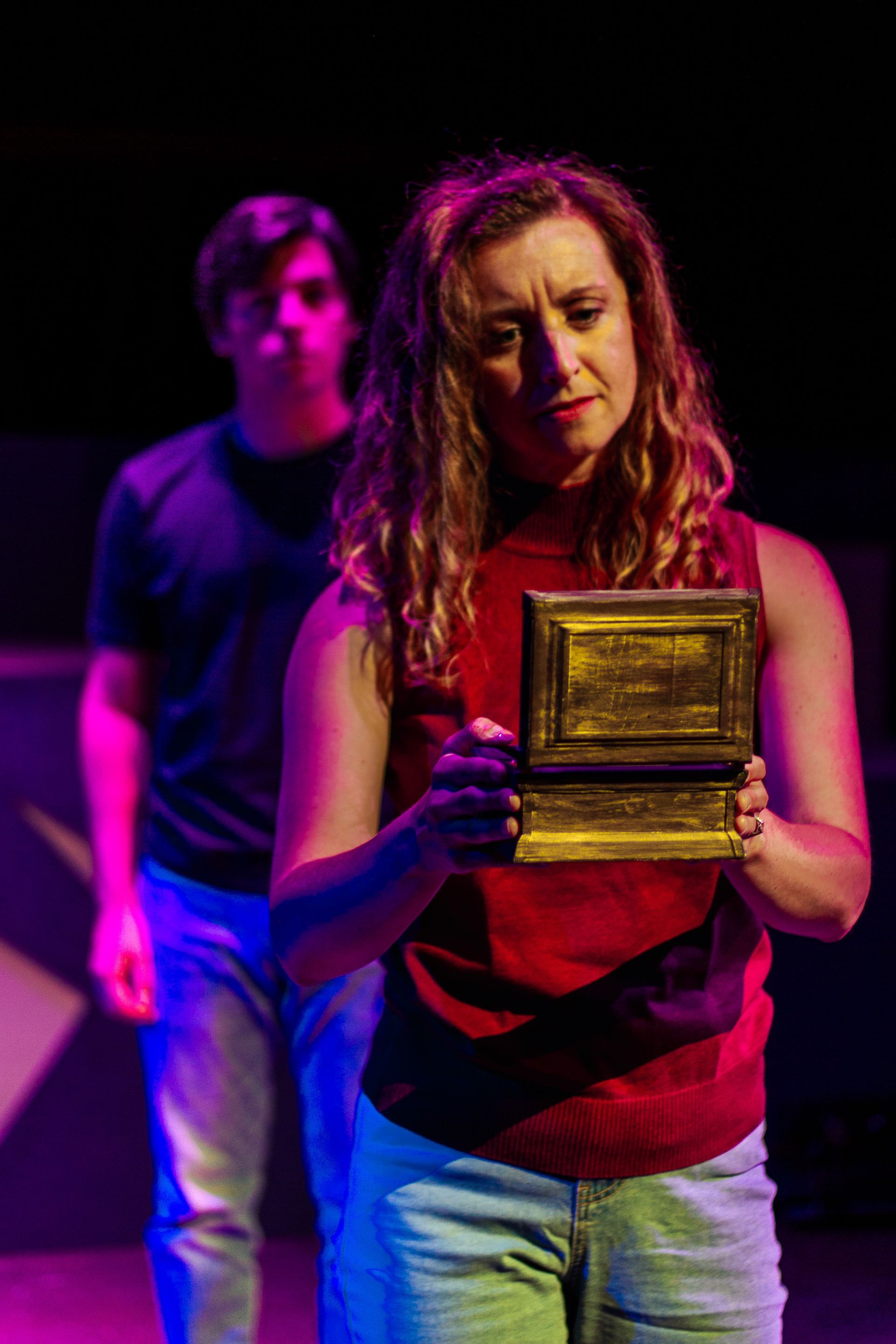Woman holding box, looking down with concern. Man stands behind, looking forward. Stage setting with purple and blue lights.