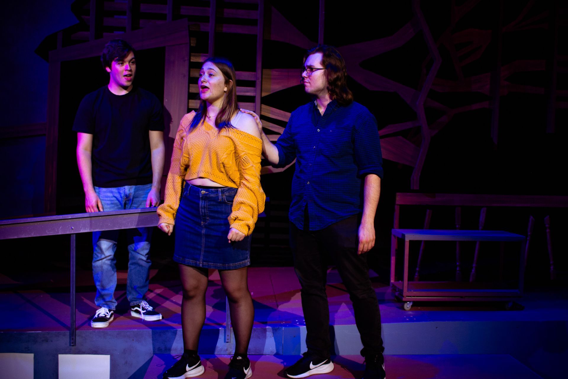 Three young people on a stage during a performance. A woman sings, men look on, dramatic lighting.