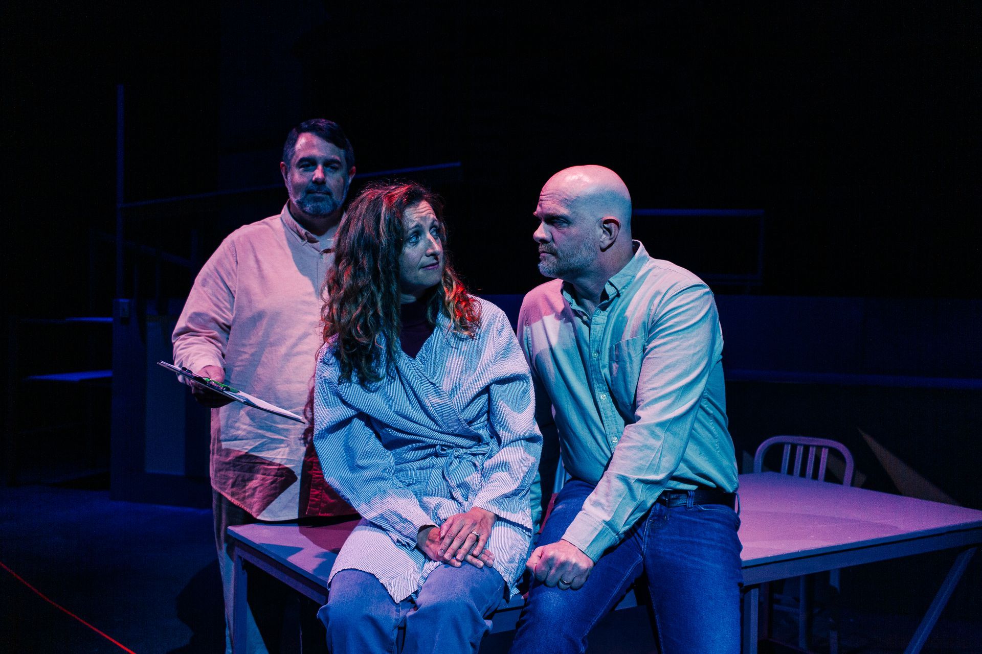 Three people on stage: a woman and bald man sit, a man stands holding a knife. Dark setting, blue light.
