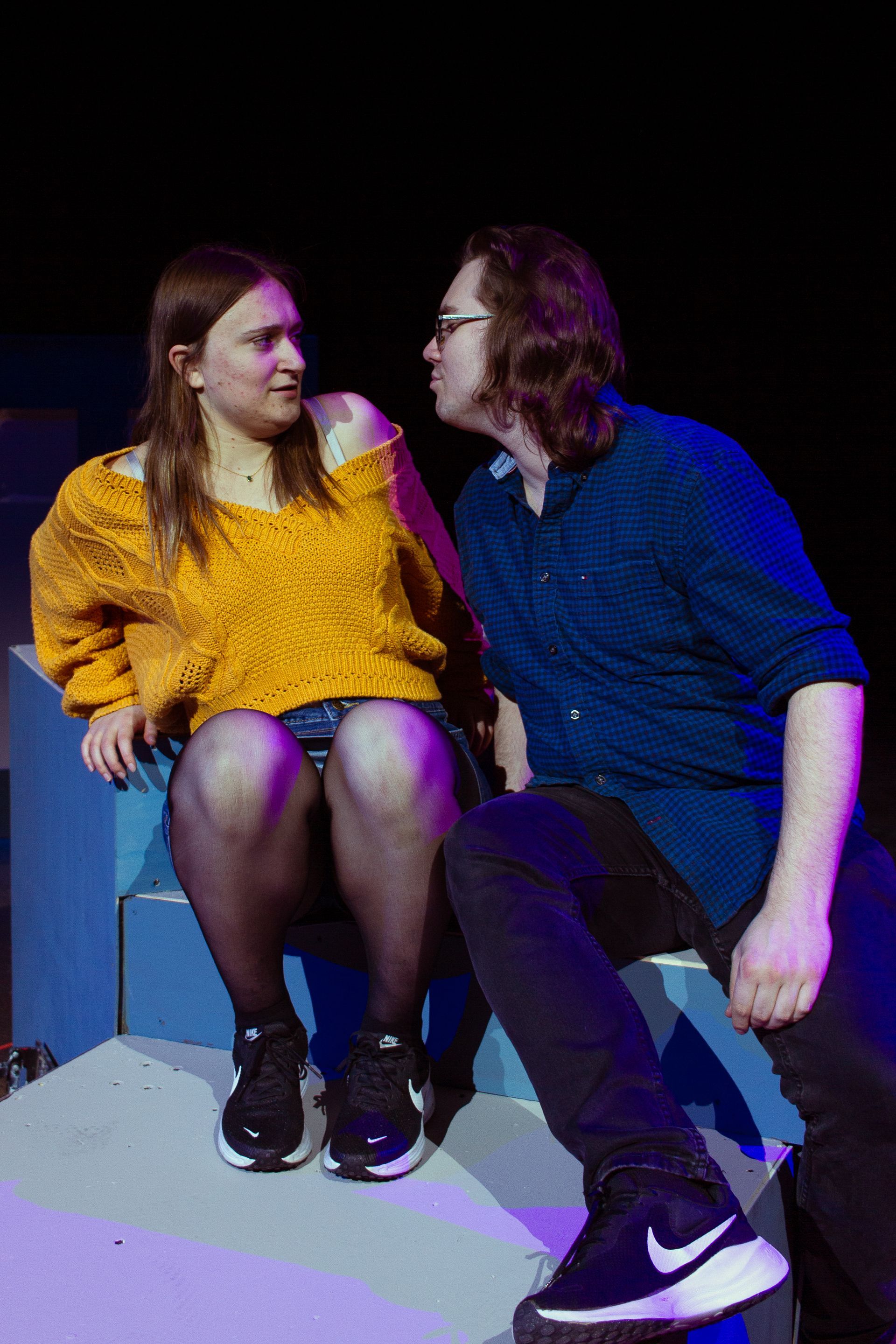 Woman in yellow sweater and man in blue shirt, interacting. They sit on blue steps; dimly lit.