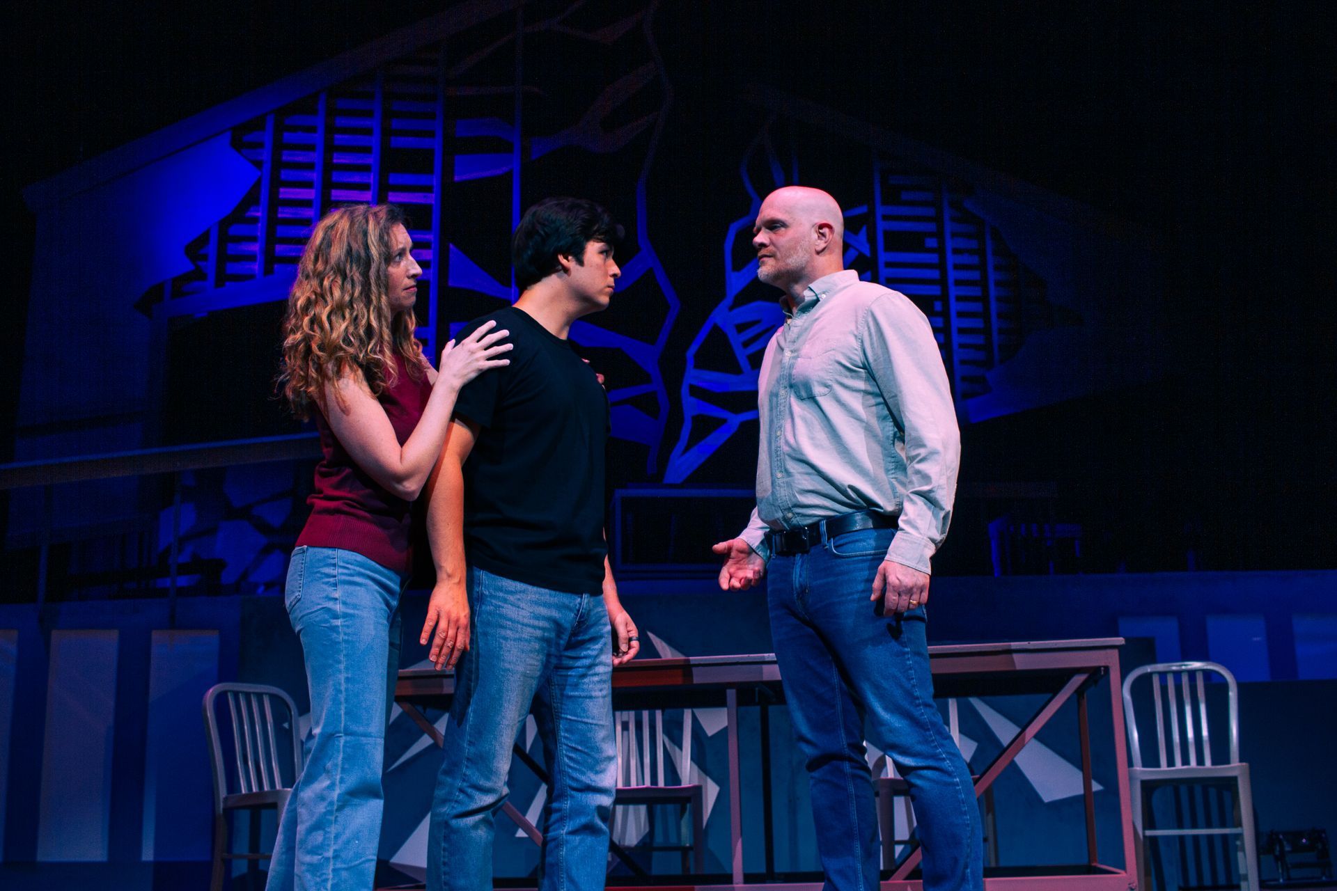 Three actors on stage: a woman with her arm around a man, facing another man in a blue-lit scene.