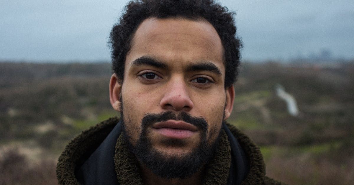 A man with dark skin and a beard, gazing at the camera with a thoughtful expression, stands outdoors.