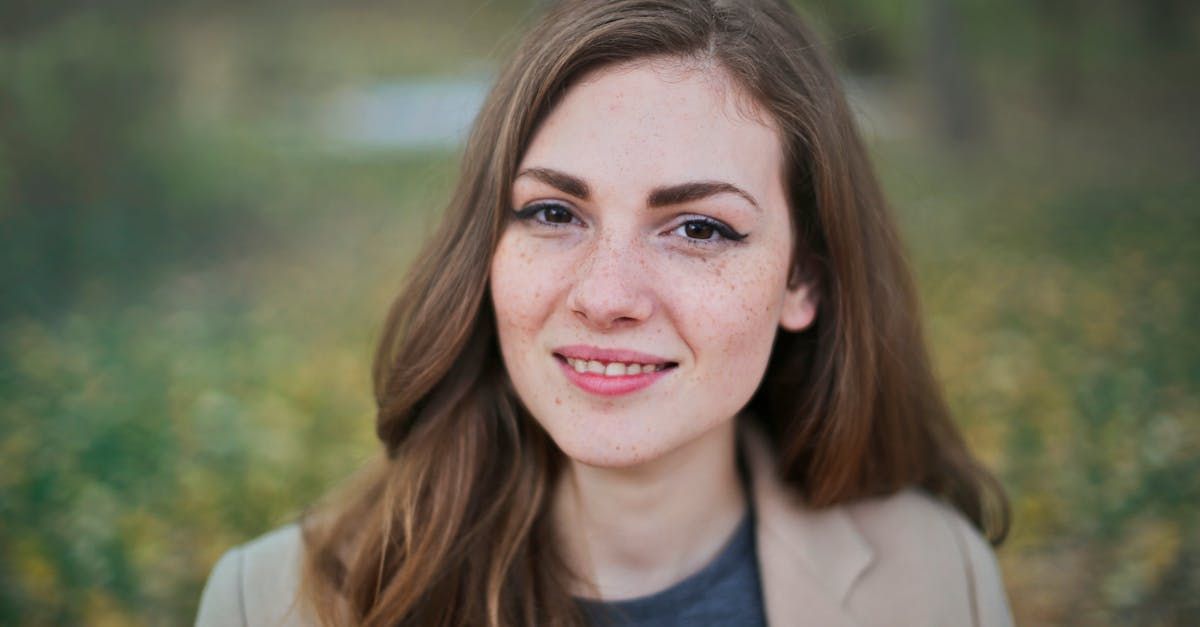 Woman with freckles smiles, wearing a tan blazer, against a blurred outdoor background.