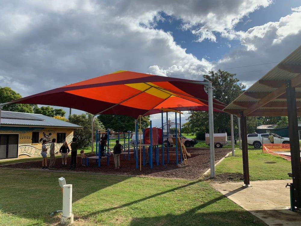 A Group Of People Are Standing Under An Orange And Yellow Umbrella In A Park — SPF Shades and Sails in Tweed Heads West, NSW