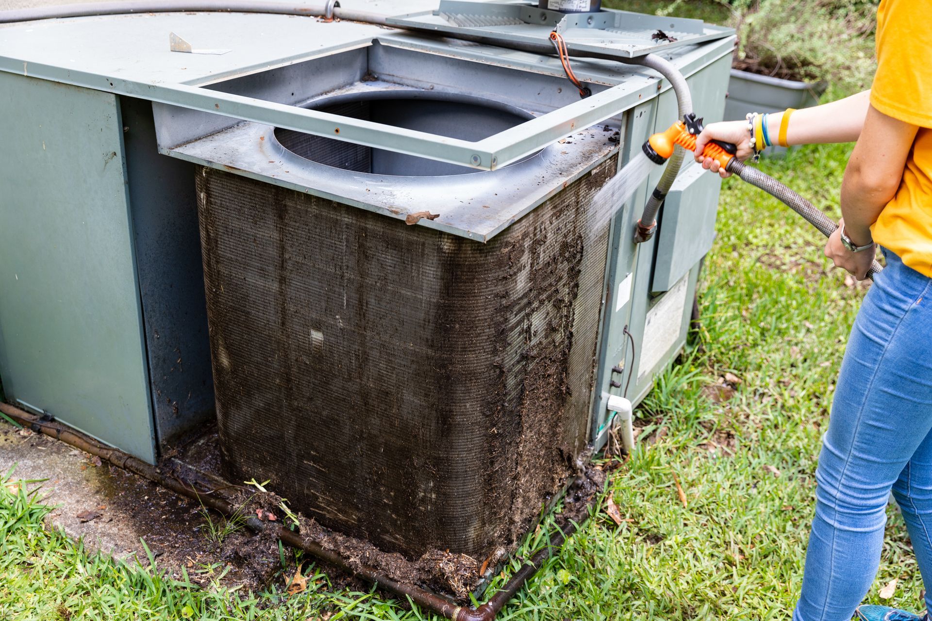 A woman is watering a dirty air conditioner with a hose.
