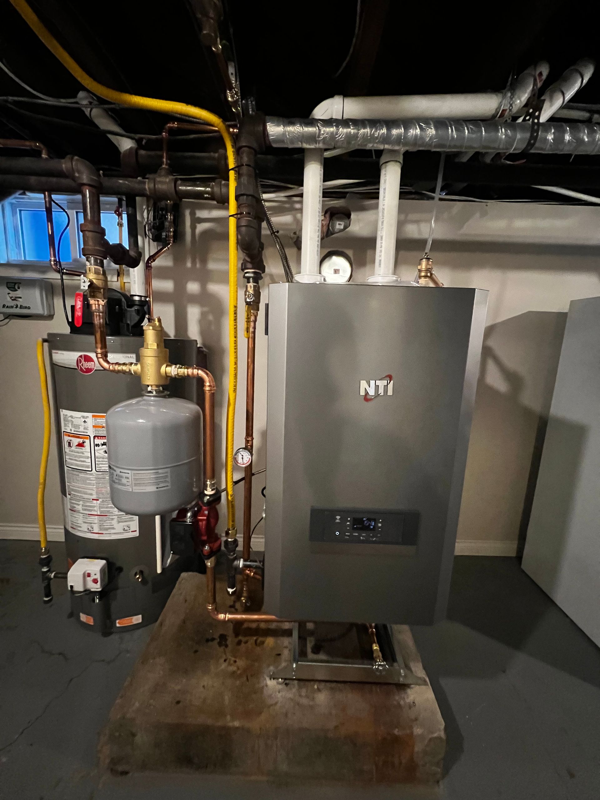 A boiler is sitting in a basement next to a water heater.