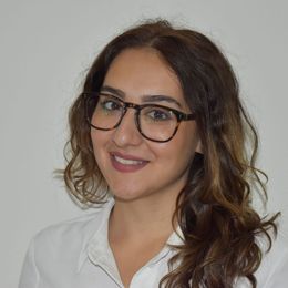 Una donna con gli occhiali, i capelli castani ricci e una camicia bianca sorride alla telecamera su uno sfondo bianco.