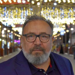 Un uomo con gli occhiali e la barba grigia, che indossa una giacca blu, è in piedi in una strada pedonale illuminata.